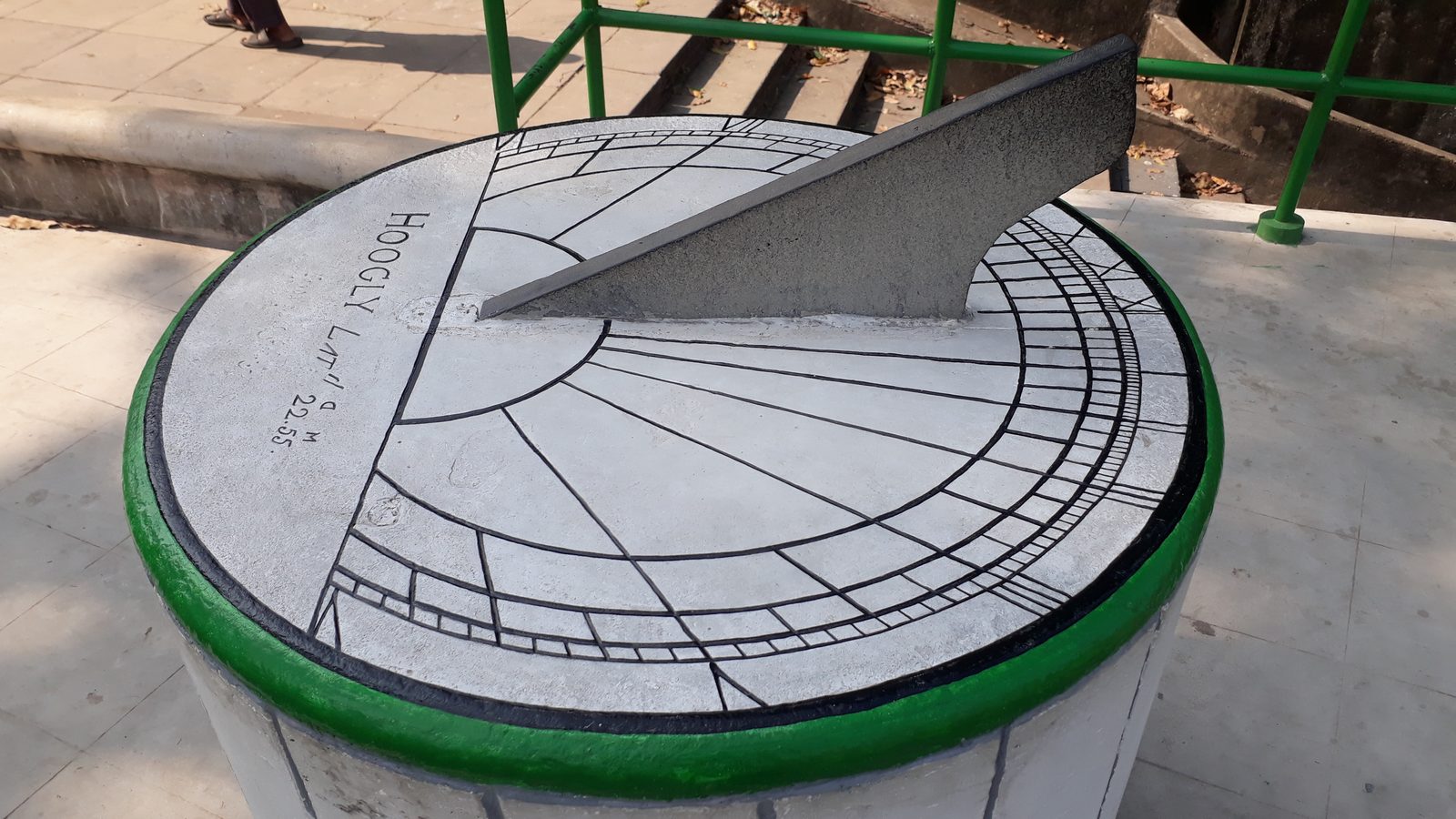 A round stone sundial standing on a low circular pillar, photographed in bright sunlight from slightly above. The pale dial face is engraved with hour lines fanning out across it and two bands of fine graduations around the rim, and a large dark triangular blade stands upright along the centre line, its edge rising towards the far side. Cut into the stone at the left, following the curve of the rim, are the words HOOGLY LAT 22.55. The pillar's rim is painted green, and a green metal railing, a flight of steps and pale paving fill the background.