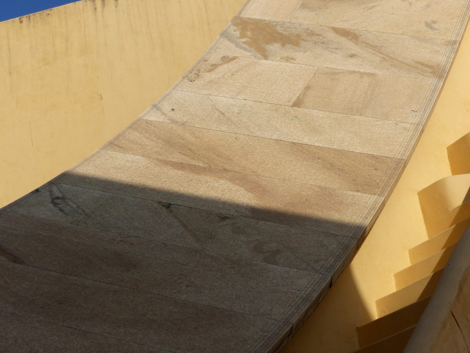 Looking down the long marble quadrant of a huge stone sundial at the Jantar Mantar in Jaipur. Fine engraved graduation lines run in bands along the outer edge of the pale marble slabs, with small numerals cut beside them. The lower half of the quadrant lies in shadow and the upper half is in full sun; the straight edge of the gnomon's shadow cuts diagonally across the scale. A yellow plaster wall and a flight of steps rise alongside, with blue sky in the top corner.