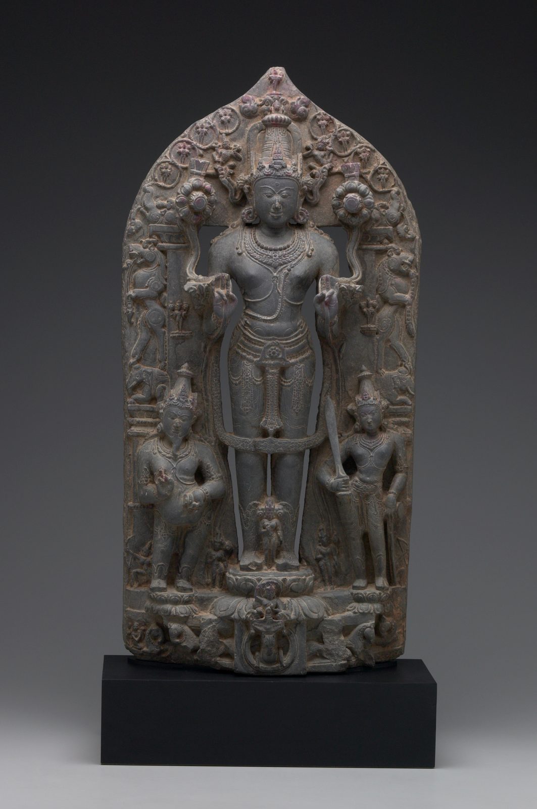 A tall panel carved in dark stone. The sun god stands upright and facing forward at the centre, holding a lotus in each hand, with small figures of his eleven brother suns carved around him on the slab. A stout bearded attendant stands at his right and a second attendant with a sword at his left, a small female figure stands between his feet, and a chariot with horses is carved along the bottom edge.