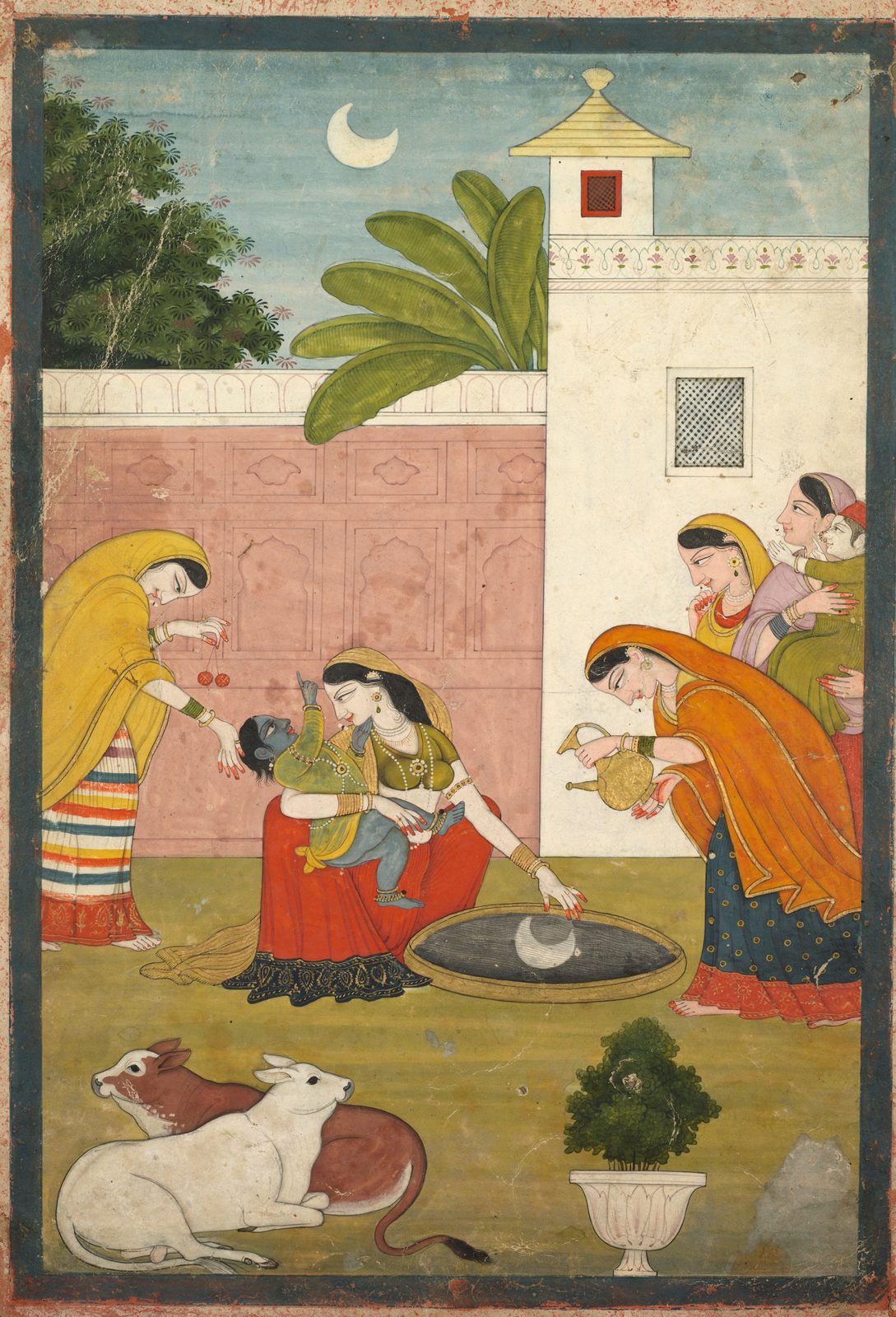 An Indian painting of a walled courtyard at dusk. A thin crescent moon hangs in the pale sky above a plantain tree and a garden wall. In the middle a woman in a red skirt sits holding the infant Krishna, who is blue-skinned and reaches out with one arm; with her other hand she points down at a wide dark dish of water on the ground, where a small white reflection of the moon is floating. A woman on the left leans in holding two round fruits, a woman on the right tips a gold jug towards the dish, and three more women stand watching. Two cows lie in the foreground beside a potted plant.