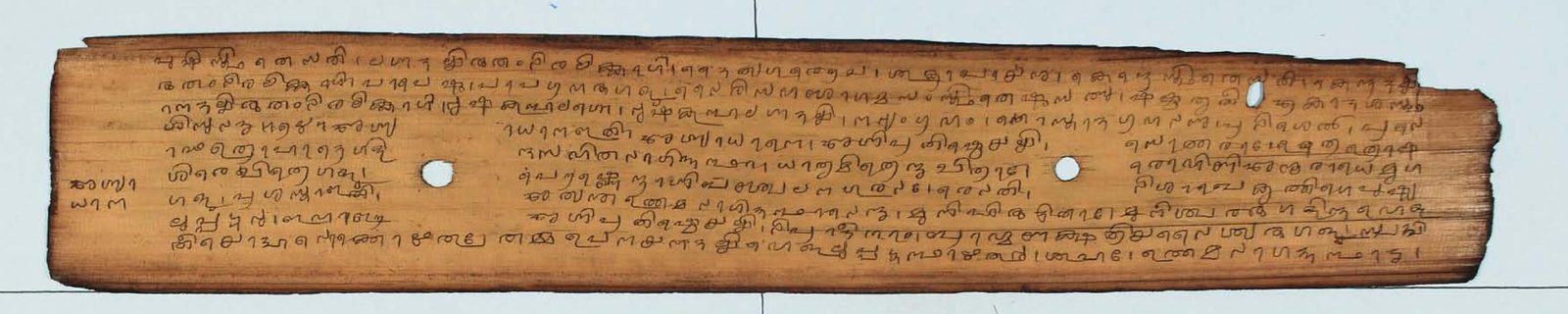 A single long palm-leaf manuscript folio lying on a pale ground. The leaf is warm honey-brown with darkened edges and is incised with nine lines of rounded Tigalari script in dark ink, interrupted by two round binding holes.