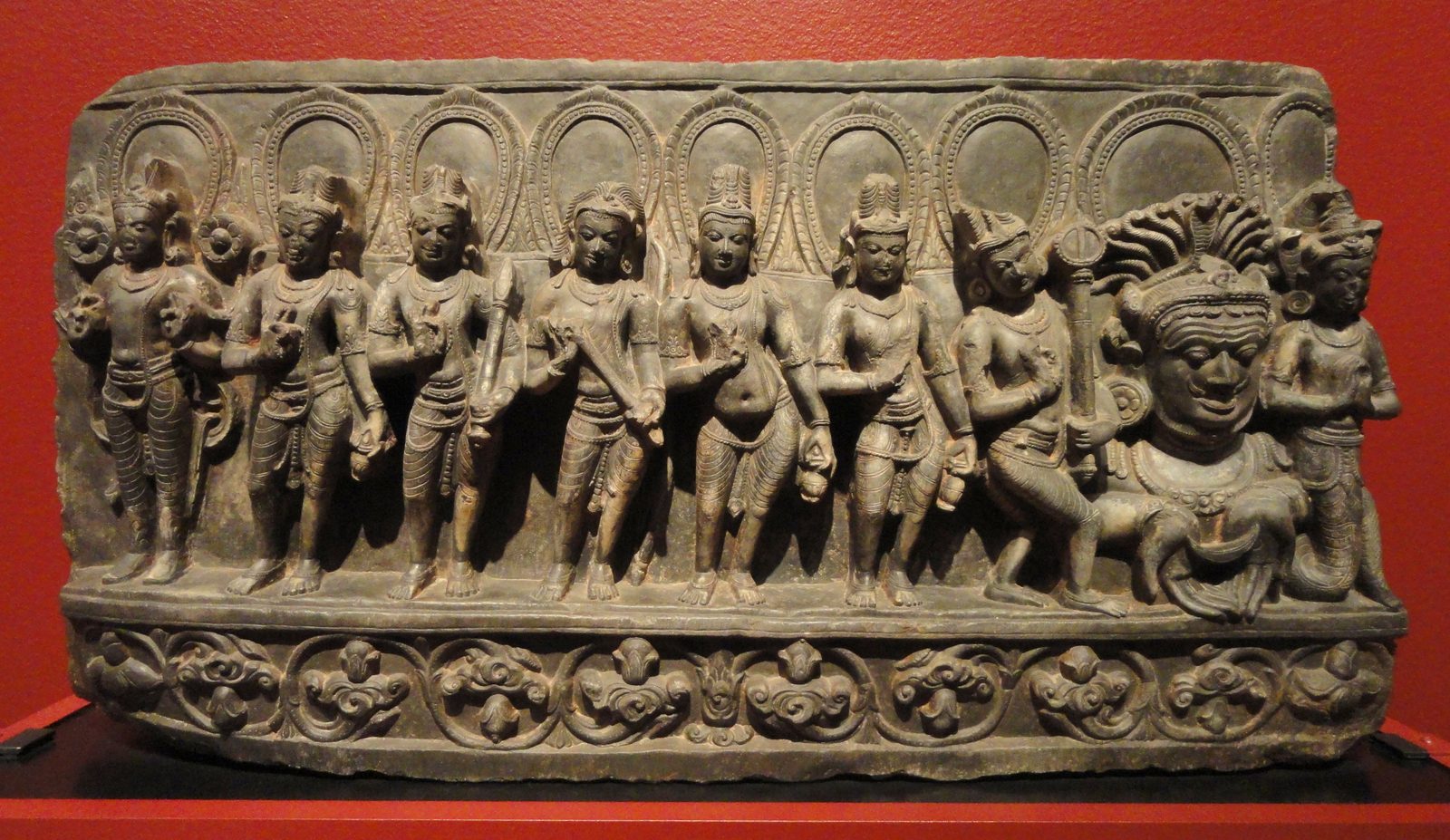 A long panel of dark grey stone, much wider than it is tall, carved with nine figures seated side by side in a single row and facing forward. They are the nine grahas of Indian astronomy, in their usual order, running from the sun at the left to Ketu at the right, and they fill the panel end to end.