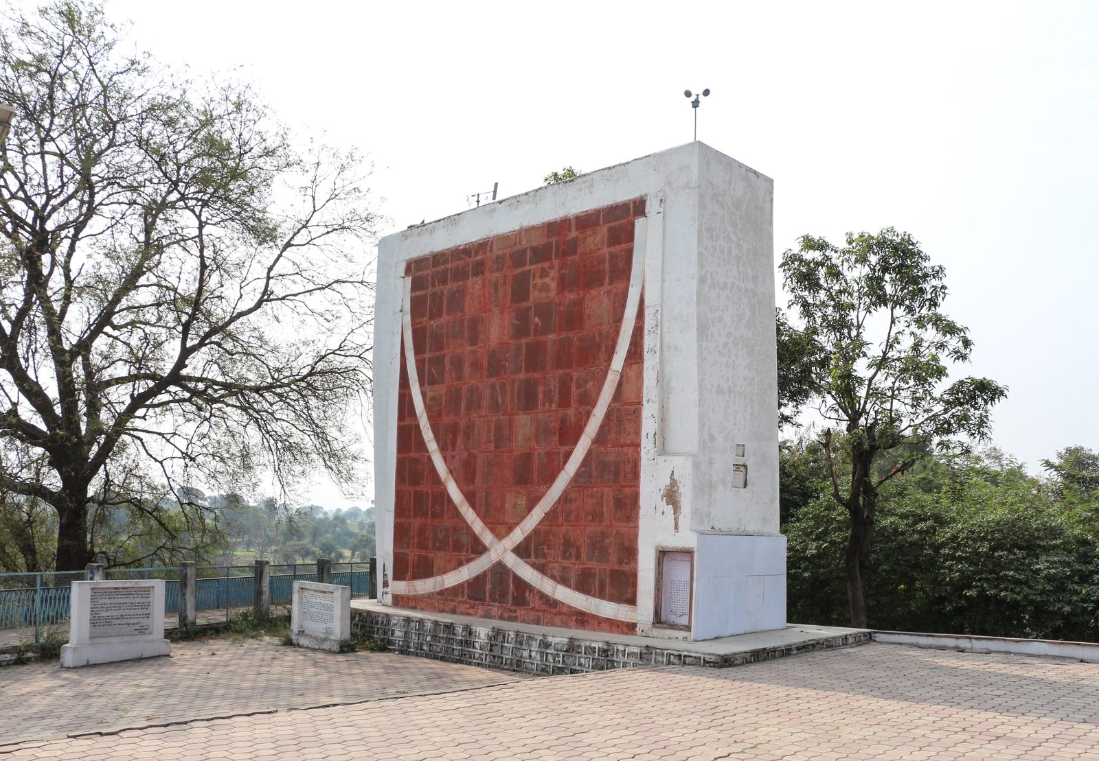 A tall rectangular masonry instrument standing alone on a paved terrace under a pale hazy sky. Its broad face is laid in dull red blocks and framed by whitewashed edges; across the lower half two white quadrant arcs sweep down and cross near the bottom centre, forming a shape like a wide bowl. The white-plastered right-hand side of the block carries a small inscribed panel near its foot. Bare trees stand to the left and leafy ones to the right, with a low blue railing and two small stone notice slabs at the base.