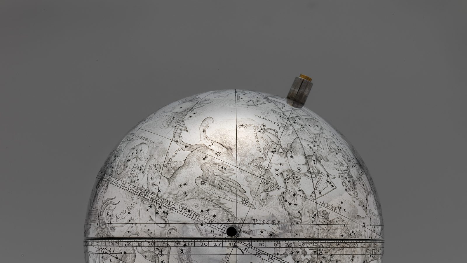 Detail photograph of the upper half of a small silver celestial globe against a plain grey ground. The polished surface is finely engraved with constellation figures, star symbols and Latin labels, among them Pisces and Aries. A graduated band crosses the globe on a slant, a ruled line runs up over the pole where a small gilt fitting projects, and a flat graduated ring marked with zodiac symbols passes across the lower part of the frame. A small dark bead sits on the slanting band where the ruled line meets it.