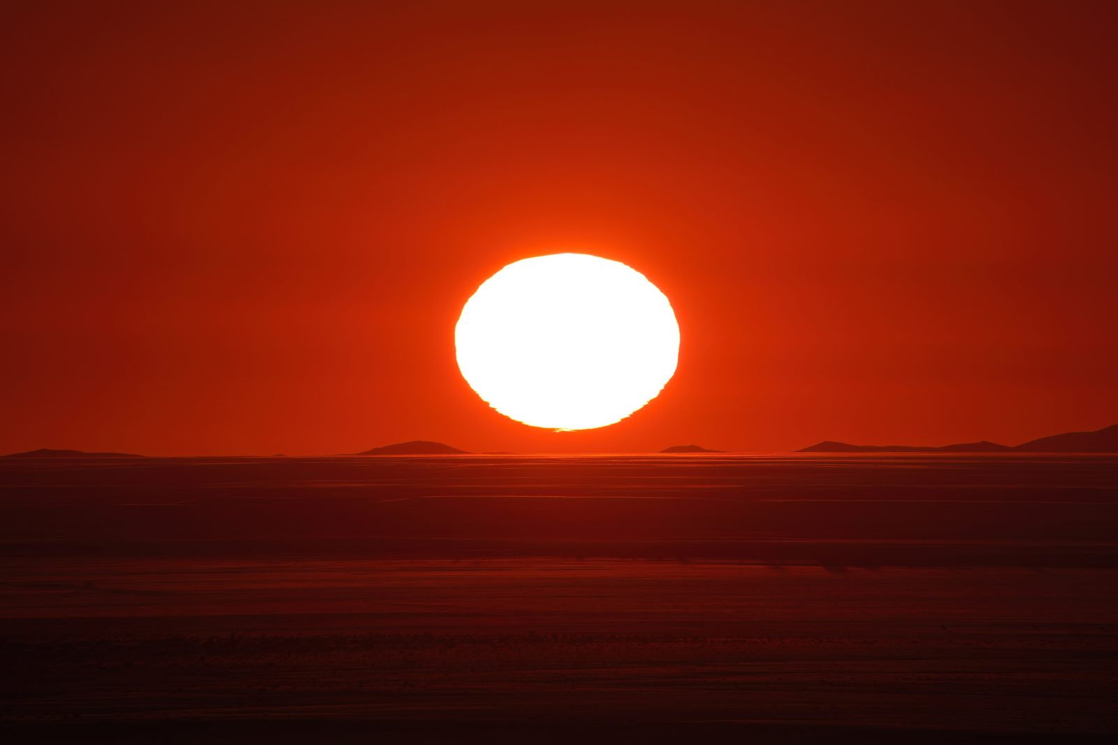 A photograph of the sun sitting on a desert horizon at the end of the day. The disc is deep orange and visibly wider than it is tall, squashed into an oval, and its lower edge is broken into ragged horizontal ripples where the air near the ground is unsteady. The land below is a dark unbroken band and the sky above shades from pale gold to grey.
