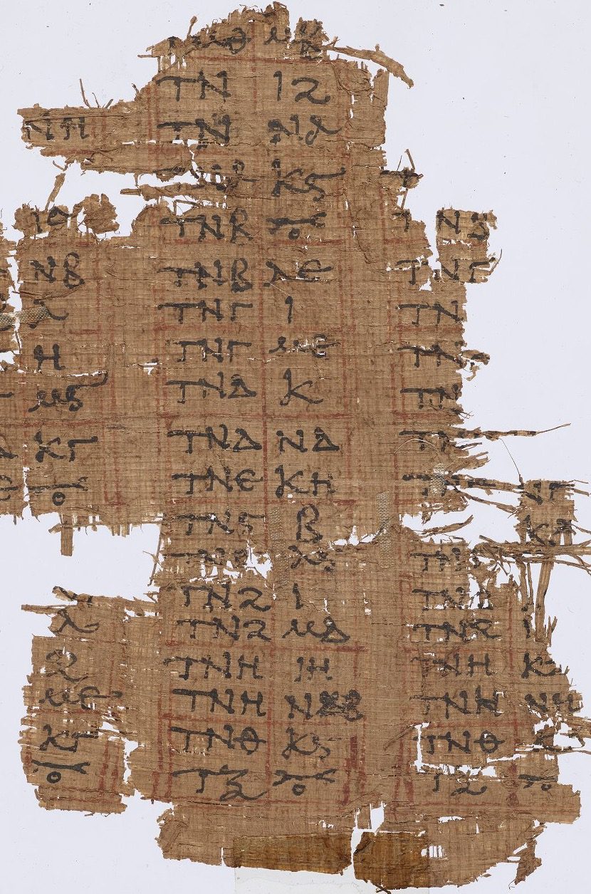 A torn fragment of brown papyrus mounted flat on a pale ground and photographed from above. Its surface is ruled into a grid by faint red lines, and each small cell holds two or three Greek letters in dark ink, the letters standing in for numerals. The columns run in short repeated groups down the fragment; several lines are broken away at the edges, where loose fibres stand out along the tears.
