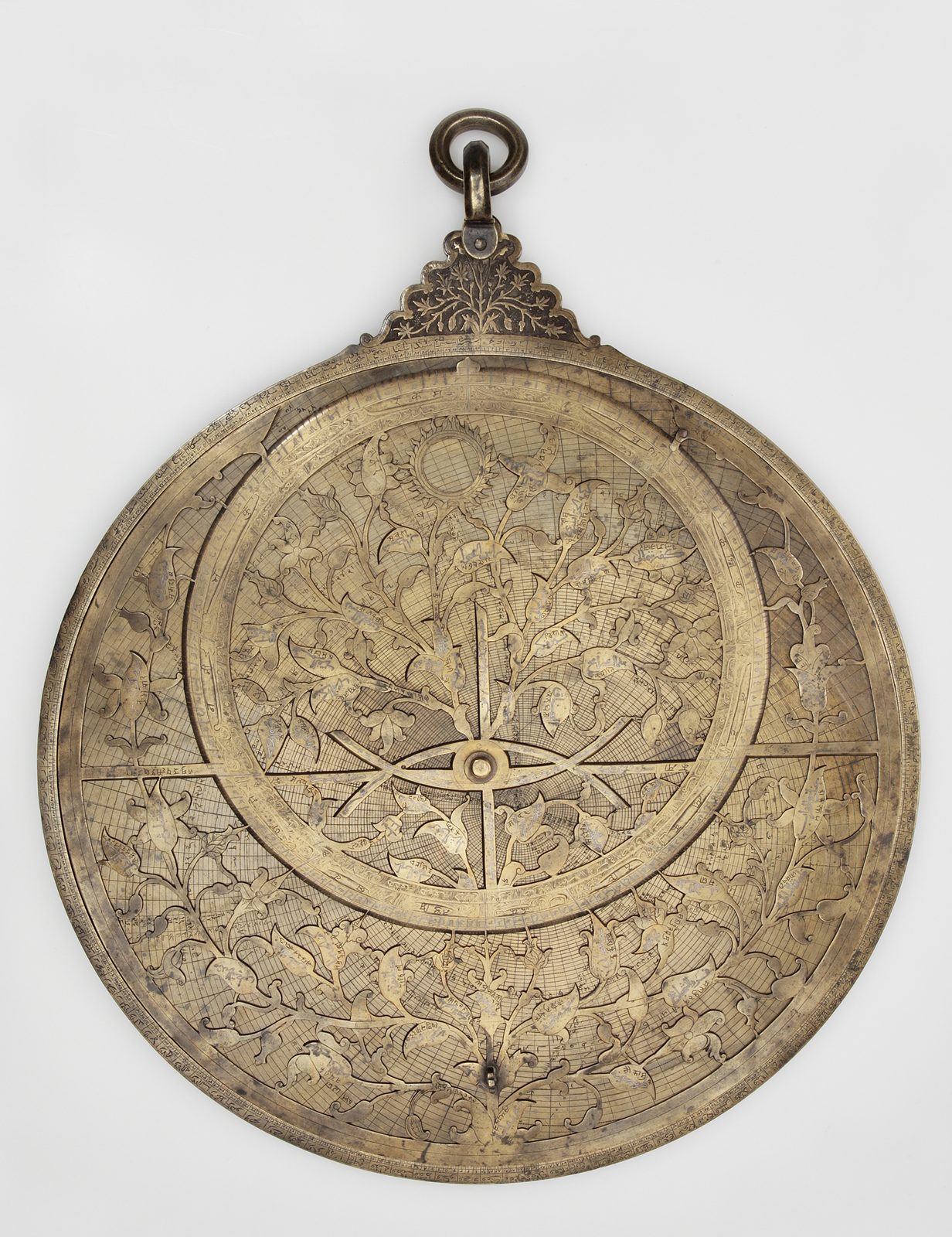 A large brass astrolabe, 52 cm across, photographed face-on against a plain pale grey ground. A pierced openwork rete of curling leaves and blossoms, each leaf tip acting as a star pointer, turns over an engraved plate covered in fine curved coordinate grids; a straight rule crosses the centre, the outer limb is graduated in degrees and finely inscribed, and an ornate leaf-cut crown with a suspension ring rises from the top.