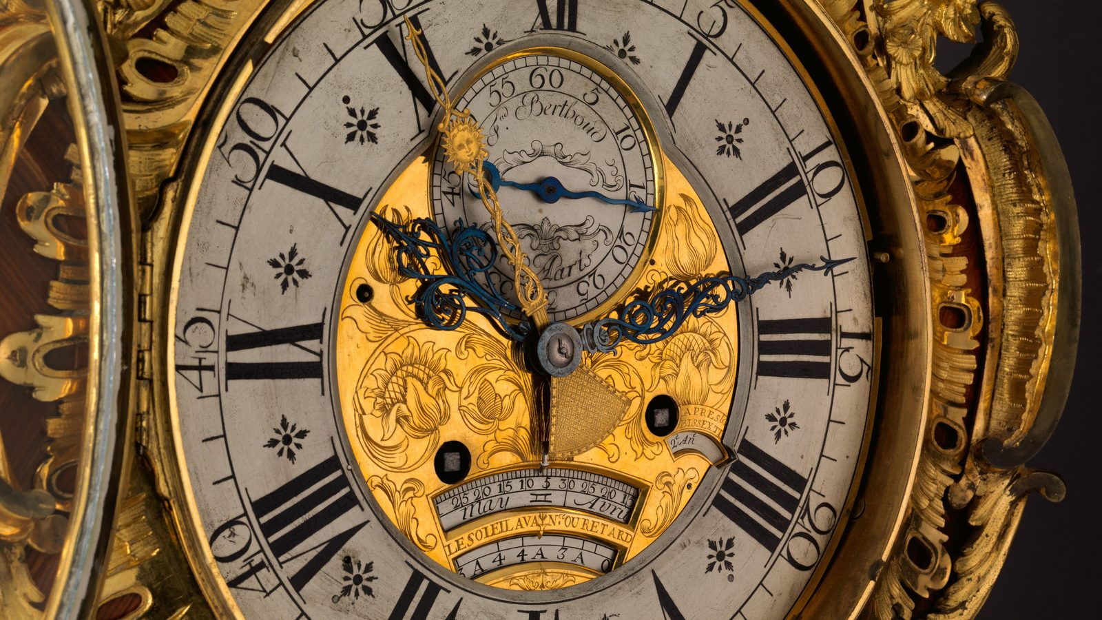 Close view of the dial of an eighteenth-century French longcase clock, seen slightly from the left. A wide silvered ring carries black Roman hour numerals with a minute ring outside it and small black florets between the numbers. The centre is a gilt plate engraved with tulips and scrolling leaves. Two blue steel hands cross it, and a slender gilt hand ends in a small sun with a face. A signed subsidiary seconds dial sits above the centre. Near the foot of the gilt plate is a small arched silvered scale marked with day numbers and the month names Avril and may, and beneath it a gilt band lettered LE SOLEIL AVANCE OU RETARDE. An ornate gilt-bronze frame surrounds the dial against a dark ground.