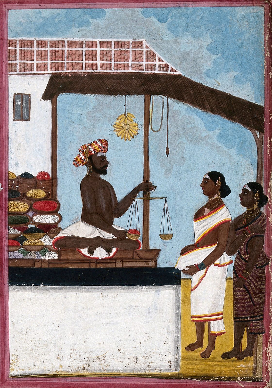 An Indian gouache painting heightened with gold, inside a pink border. Under a thatched awning at the front of a tiled-roof shop, a seller sits cross-legged on his stall in a white dhoti and a tall red and yellow patterned turban, with large gold hoops in his ears. He holds a small two-pan balance up by its beam, one pan hanging lower than the other, and steadies a shallow dish of something red on the stall with his free hand. Open dishes and baskets of grains, pulses and sweets in yellow, red, green, white and black are ranged along the stall beside him, and a bunch of yellow bananas hangs from the awning overhead. Two women in white and striped cloth, wearing gold necklaces and bangles, stand on the sandy ground at the right, waiting while he weighs.
