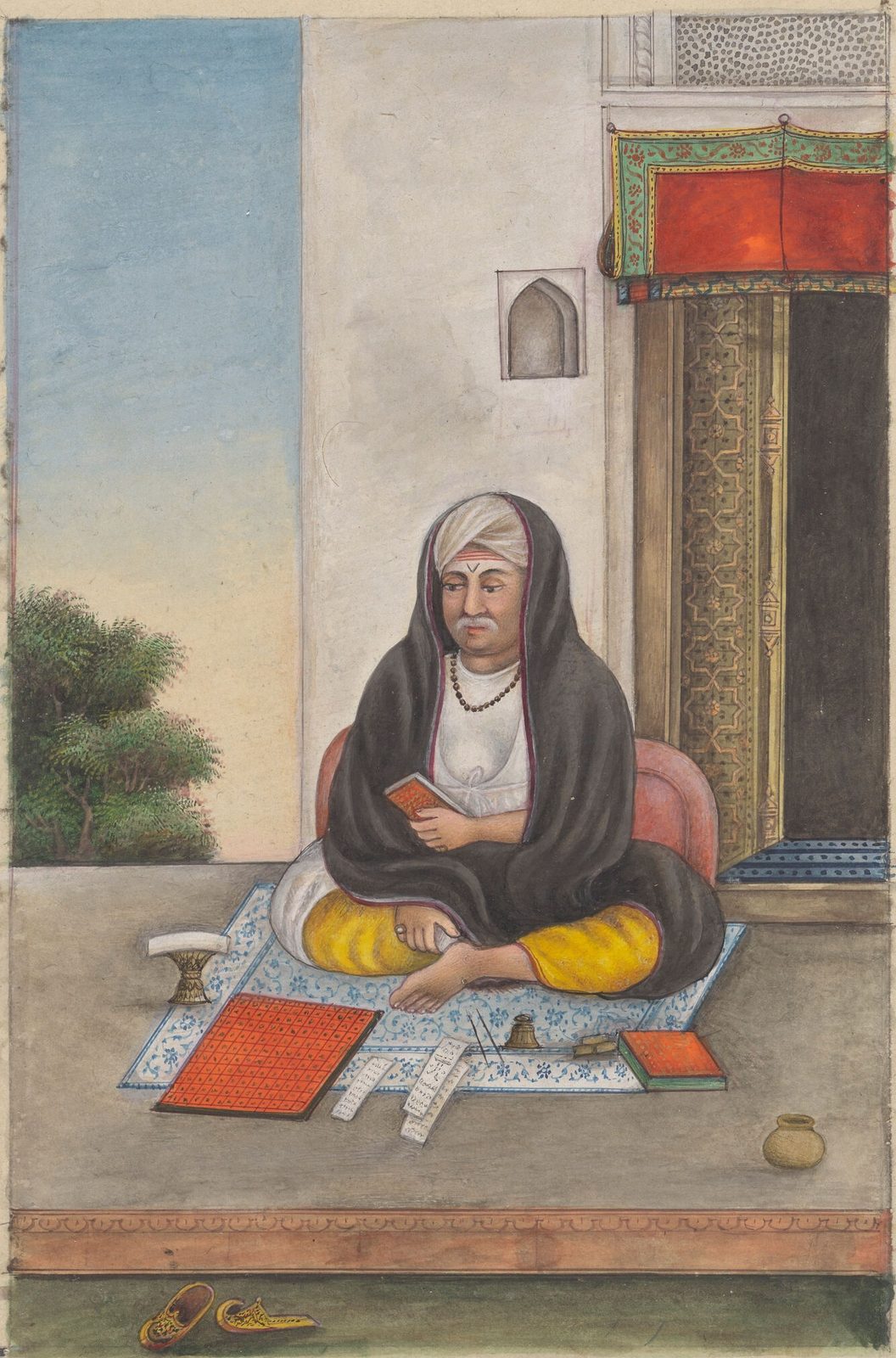 A Company-school painting on aged cream paper. A Brahmin astrologer sits cross-legged on a blue-and-white patterned mat laid on a stone step outside a doorway, wrapped in a dark shawl over white, a white turban with a red caste mark on his forehead, a small red-bound book held in one hand. Spread on the mat in front of him are a red chart ruled into a grid of squares, a folded paper manuscript covered in columns of writing, an inkpot with a pen, a second red-bound book and a small brass water pot. His slippers lie on the ground below the step. A green tree and pale sky show at the left; a carved wooden door with a red awning stands at the right.