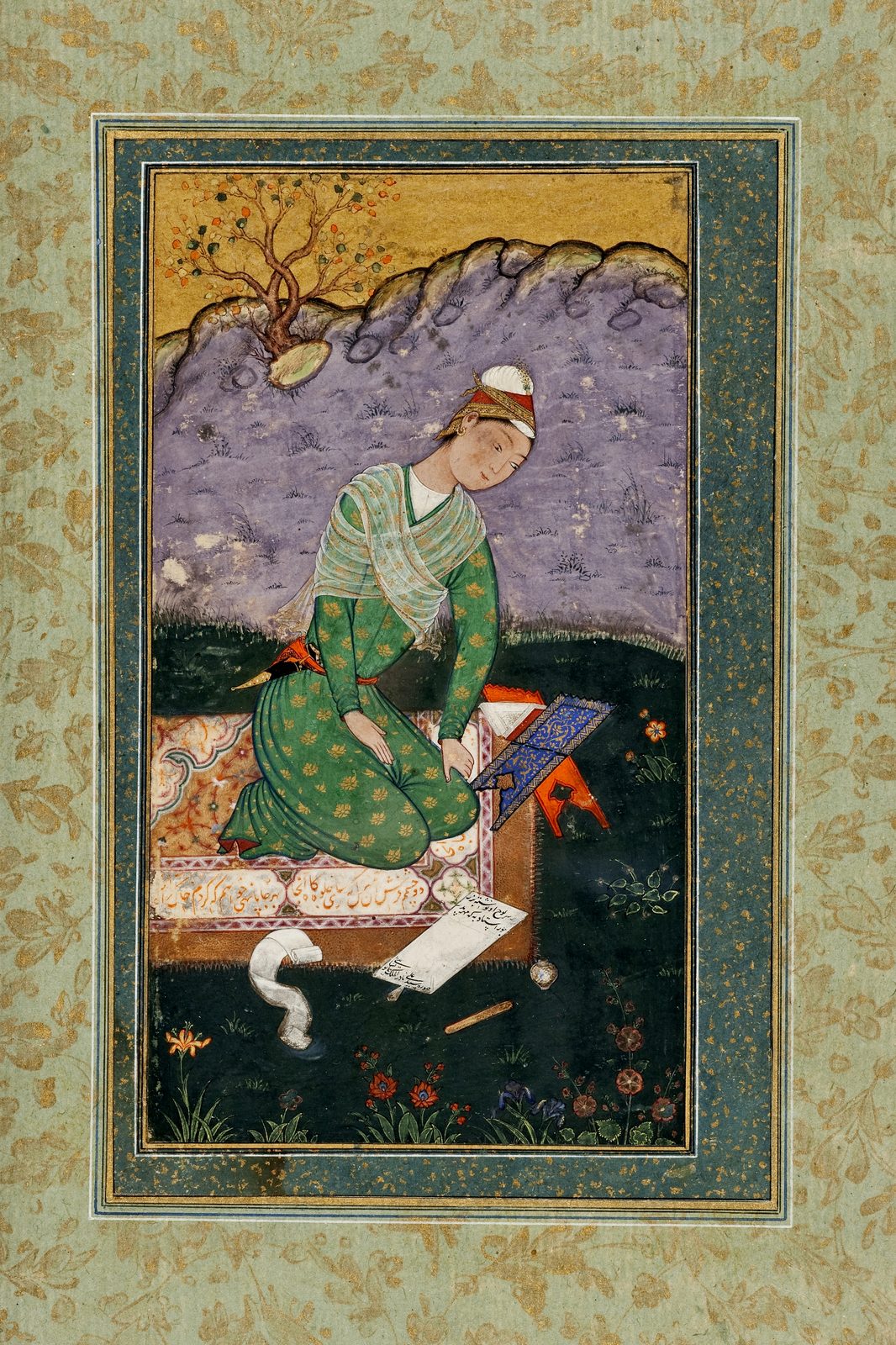 A Mughal album page. A young man in a green robe patterned with small gold flowers kneels on a rug spread over dark green ground, a pale striped shawl across one shoulder and a white turban wound over a red and gold cap. He leans forward with both hands on his knees, looking down at an open book propped on a low folding stand of orange lacquer whose cover is worked in blue and gold. On the ground in front of him lie a part-unrolled white scroll, a flat sheet covered with lines of Persian writing, a small white inkpot and a reed pen. Behind him a lavender hillside rises against a gold sky with one flowering tree; small red and blue flowers grow along the lower edge. The painting sits inside ruled gold borders on a pale green album margin sprinkled with gold flowers.
