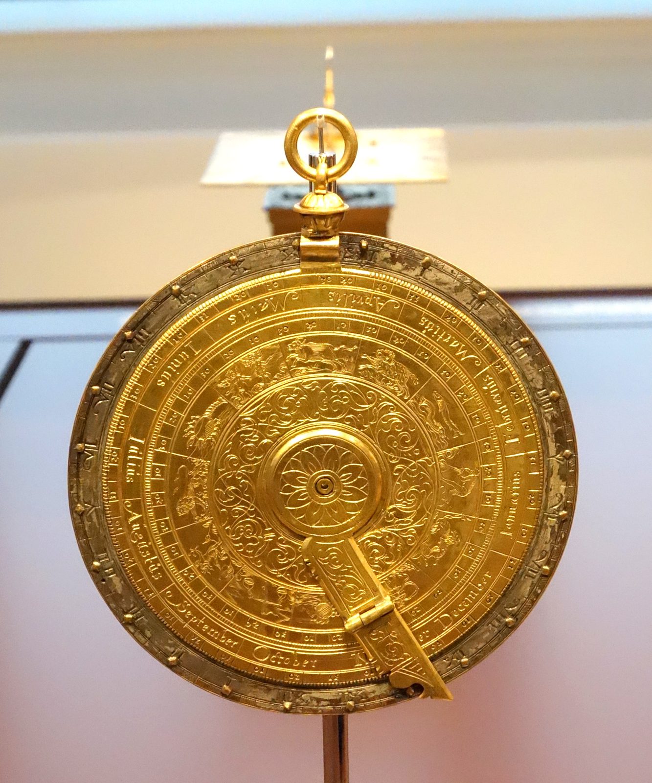 A brass nocturnal made in Prague around 1600, photographed in a museum. It is a flat disc mounted on a long straight handle. Rings around the disc carry engraved scales and small numerals, a hole is pierced through the centre, and a slim arm is pivoted at that centre so that it can be swung round the face like the hand of a clock.