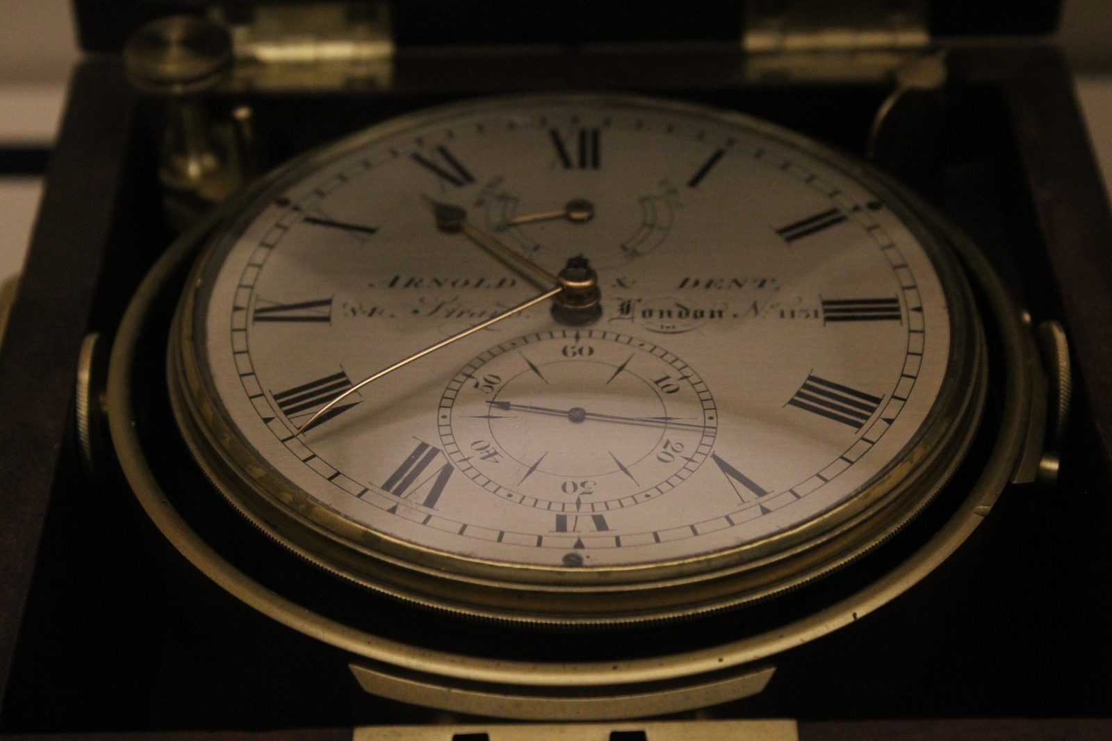 A marine chronometer in its fitted wooden case with the lid open, photographed from slightly above. The round white dial carries numerals and a small separate seconds dial, and the instrument is held in a brass ring so that it stays level when the case is tilted.