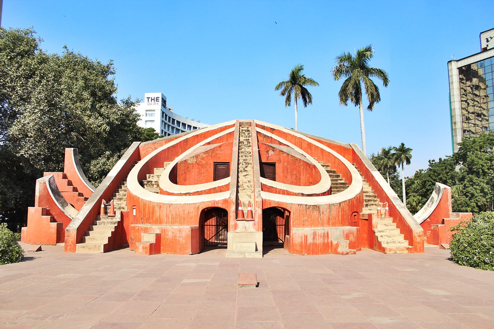 The Misra Yantra at the Jantar Mantar observatory in Delhi, a large masonry instrument standing in the open air. A central triangular gnomon wall rises between two broad curved quadrant faces, with further walls and flights of steps set around it, all of it built in plastered stone in the deep red of the Delhi site, and graduated scales marked along the curved surfaces.