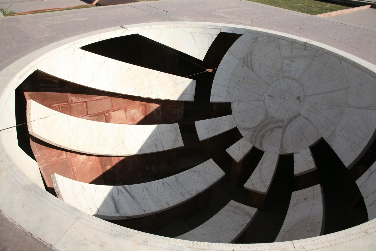Looking down into one of the Jai Prakash Yantra bowls at Jaipur. A hemisphere sunk into the ground is lined with curved white marble segments, each finely engraved with a grid of coordinate lines and graduated scales; deep gaps between the segments open onto red sandstone walls and dark shadow below. Thin cross-wires are stretched across the mouth of the bowl, and a hard diagonal shadow falls across the left half.