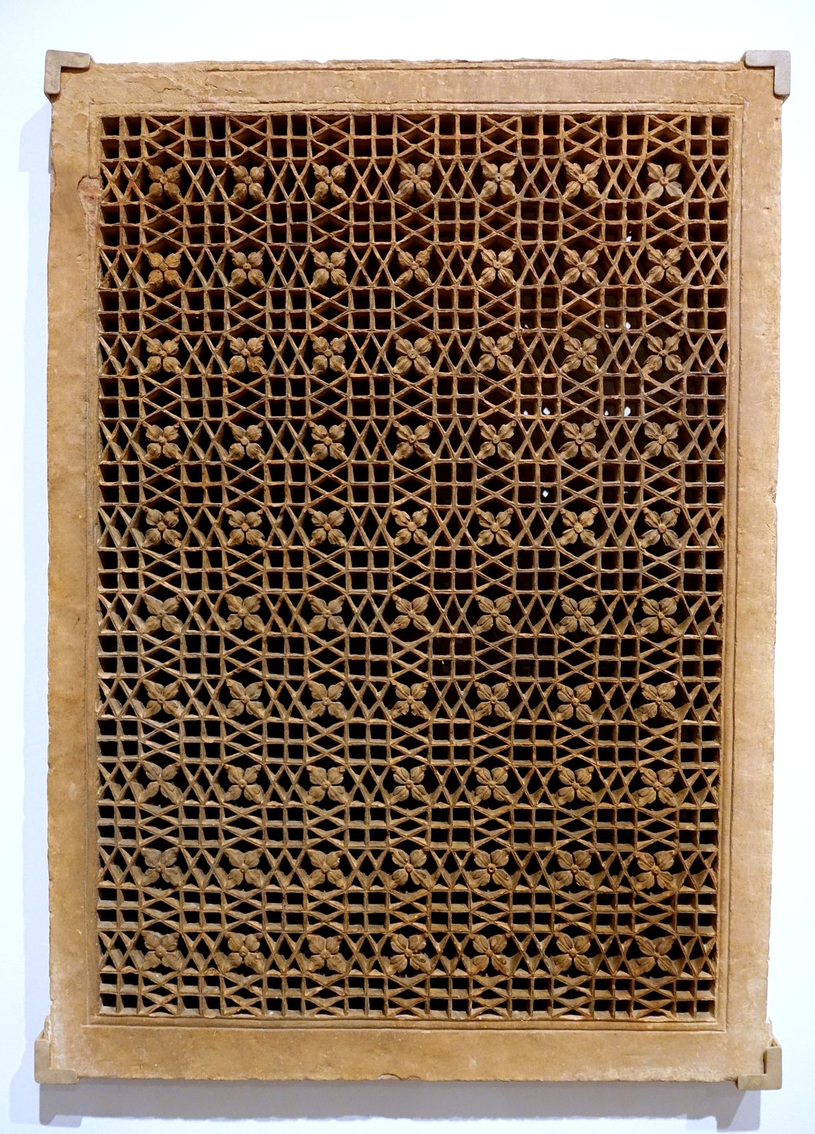 A pierced stone screen from Mughal India, photographed against a plain museum wall. The sandstone panel is cut right through into a close repeating lattice, so that the light and the wall behind show through every opening.