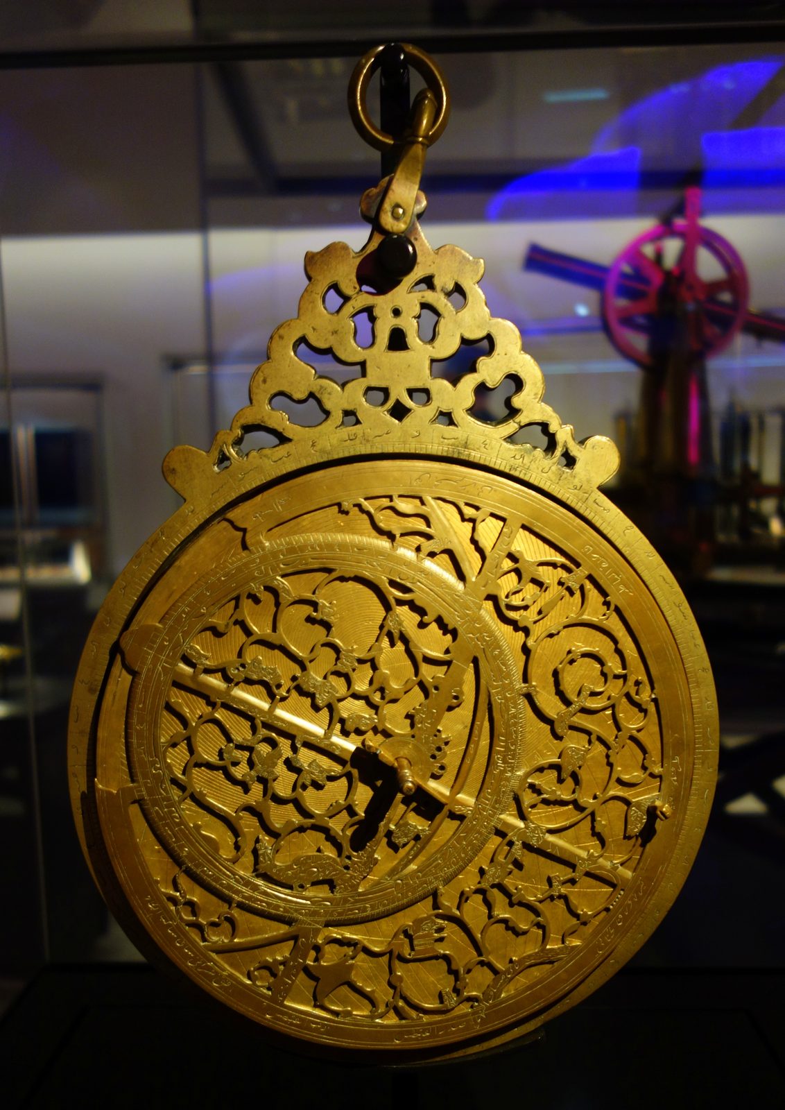 A brass astrolabe hanging by its ring in a museum case, photographed from the front. Above the disc sits a pierced openwork crown. The face is a fretwork of curling leaf-shaped pointers over engraved circles, with a straight rule laid across the middle, and a band of Arabic script and graduated marks runs round the rim. The metal is warm gold against a dark background.