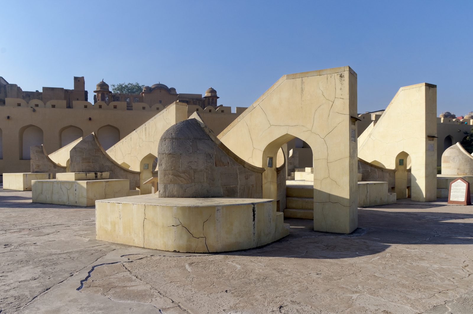 The Rashivalaya Yantra at the Jantar Mantar observatory in Jaipur: a group of twelve small stone instruments, one for each sign of the zodiac, standing side by side on an open paved terrace. Each is built to the same plan, a triangular gnomon wall of pale masonry rising at an angle from a curved quadrant face marked with graduated scales.