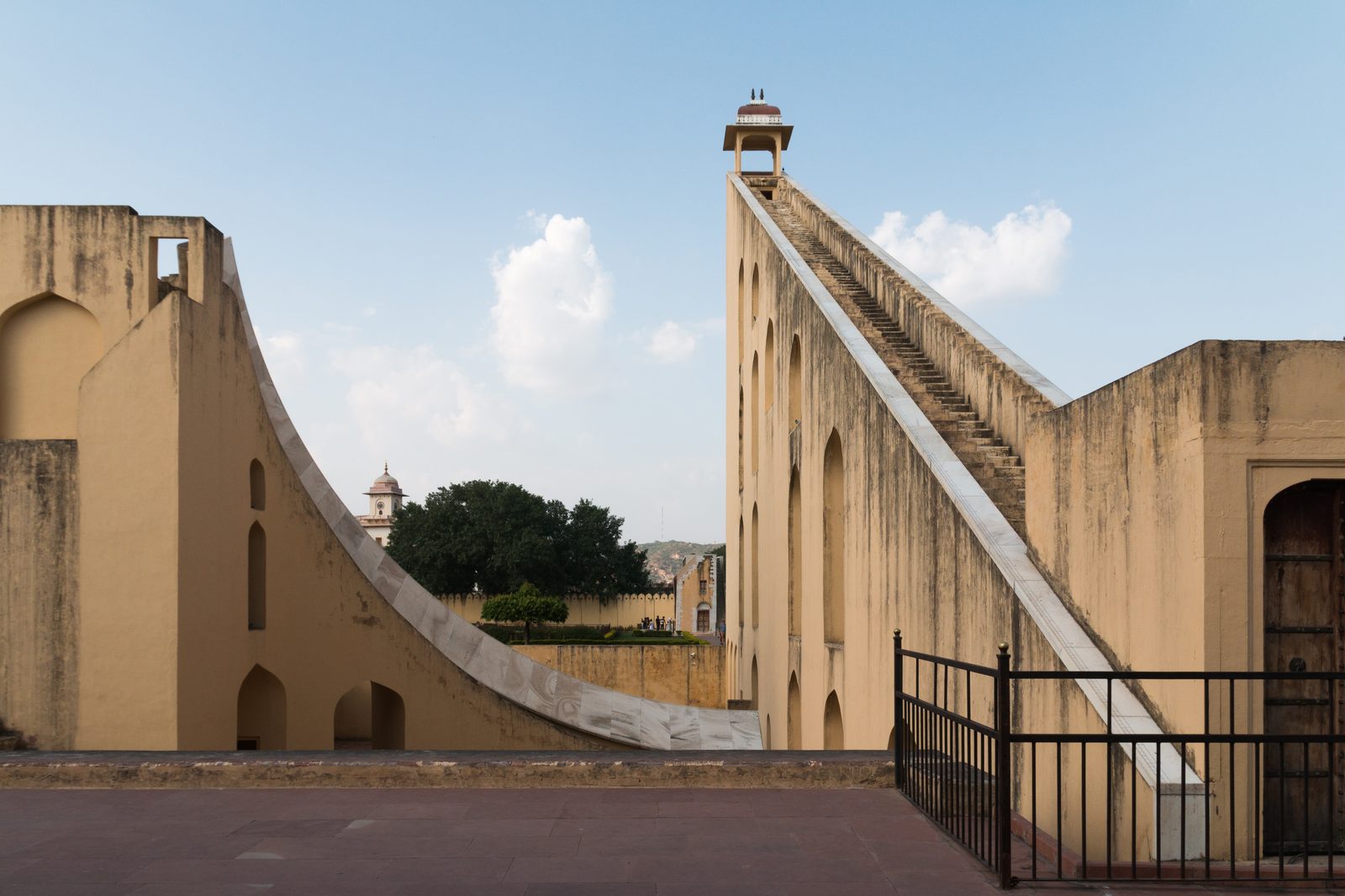 The Brihat Samrat Yantra at the Jantar Mantar observatory in Jaipur, a sundial the size of a building. On the right a steep triangular gnomon wall rises to a small domed pavilion, its sloping edge faced with a marble stair; on the left a curved marble-faced quadrant sweeps down towards the paved ground. Warm cream masonry against a blue sky with one white cloud.