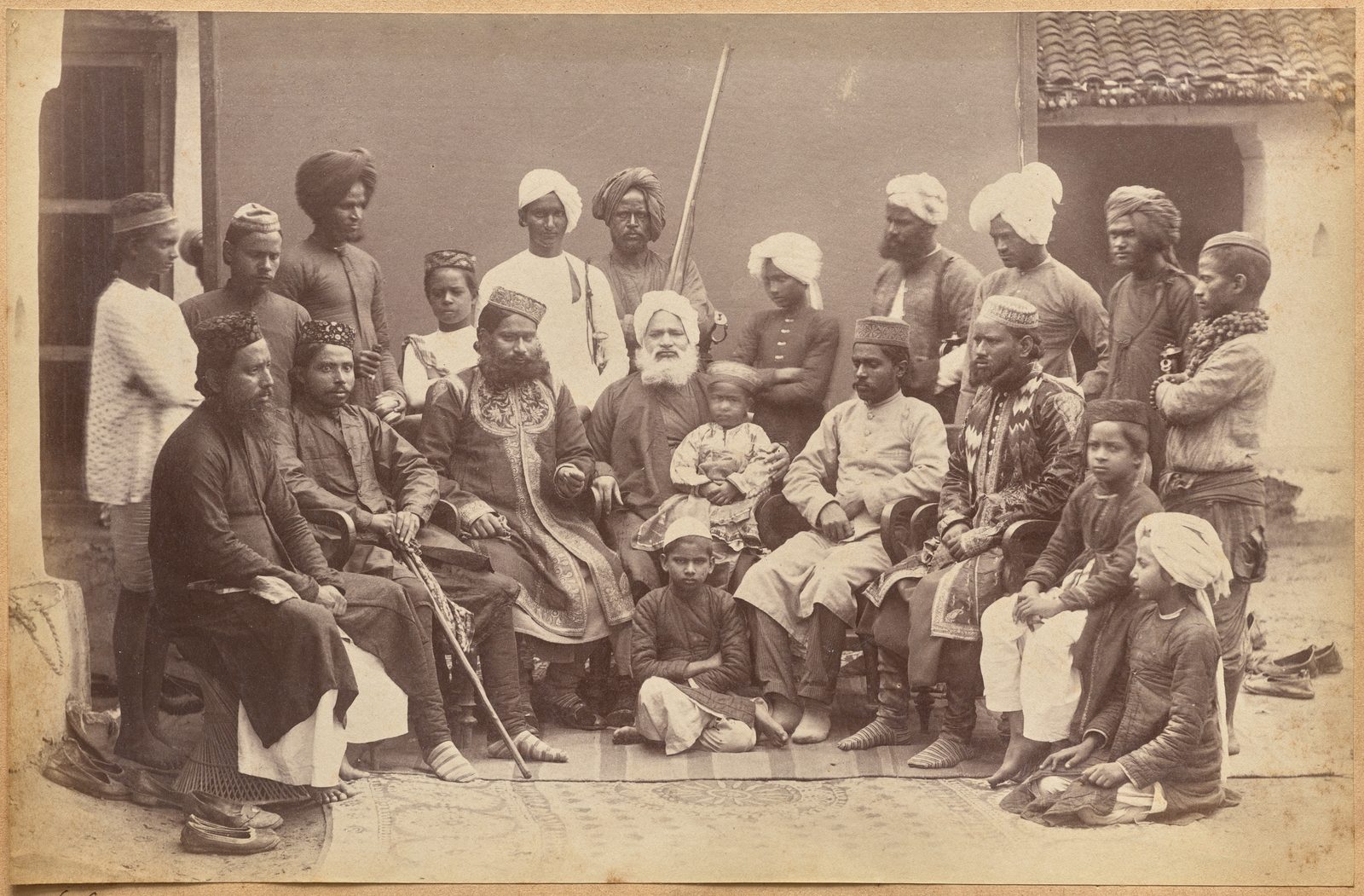 A sepia albumen photograph on a browned album mount, wider than it is tall. About twenty men and boys of one household are grouped outside a plastered house with a tiled roof. Eight sit on chairs at the front, on a patterned carpet laid over bare ground: bearded men in dark coats and embroidered caps, one resting both hands on a walking stick, one in a heavily embroidered robe, and an elderly man with a full white beard holding a small child on his lap. A boy sits cross-legged on the carpet in front of them and two more boys sit at the right edge. Behind the chairs stand a dozen younger men and attendants in turbans and white caps, one with a sheathed sword at his side and one holding a tall pole.