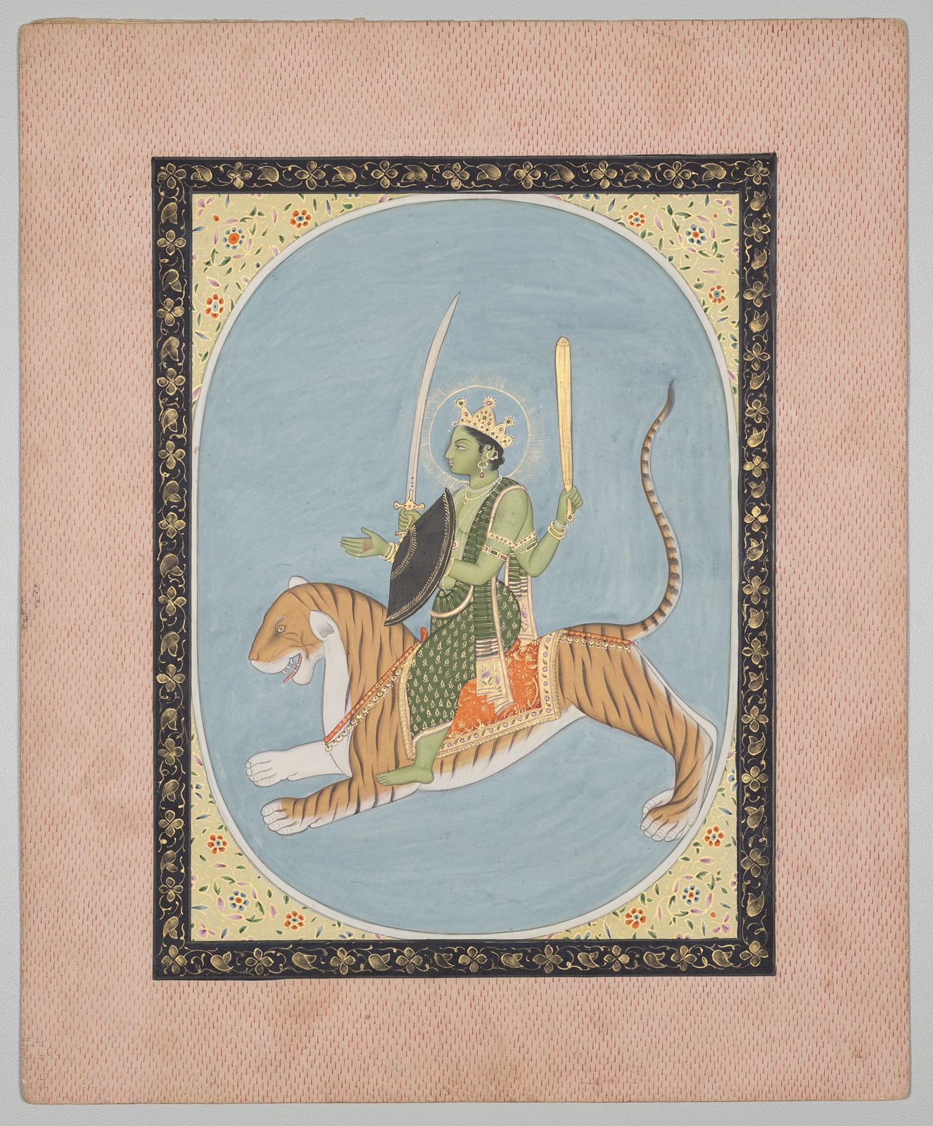 An Indian miniature painting of the planetary deity Mercury inside a pale blue oval. A green-skinned figure with four arms, a gold crown and a gold halo rides a leaping orange tiger, holding a curved sword, a shield and a gold club. The oval is set in a border of yellow and black floral panels within a textured pink margin.