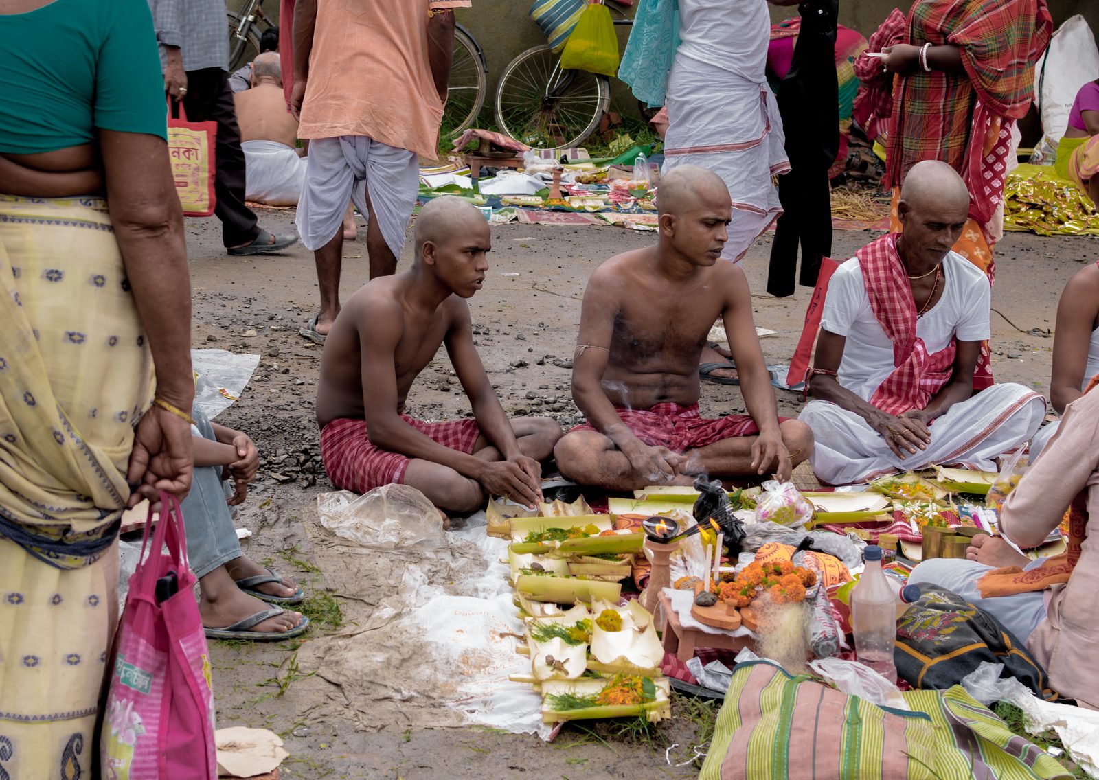 Men in white cloth stand in shallow river water in the early morning, hands raised together, making offerings of water to their ancestors while a priest conducts the rite from the bank.