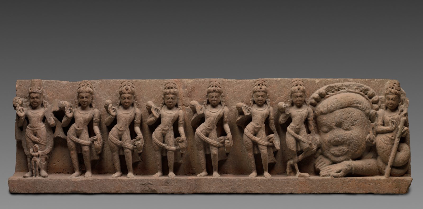 A long, weathered sandstone temple lintel carved with nine figures in a single row: seven standing crowned men holding small water pots, then a large disembodied head with a mane of hair and two hands resting on the ledge, and at the far right a standing figure with a staff whose lower body coils into a serpent's tail.