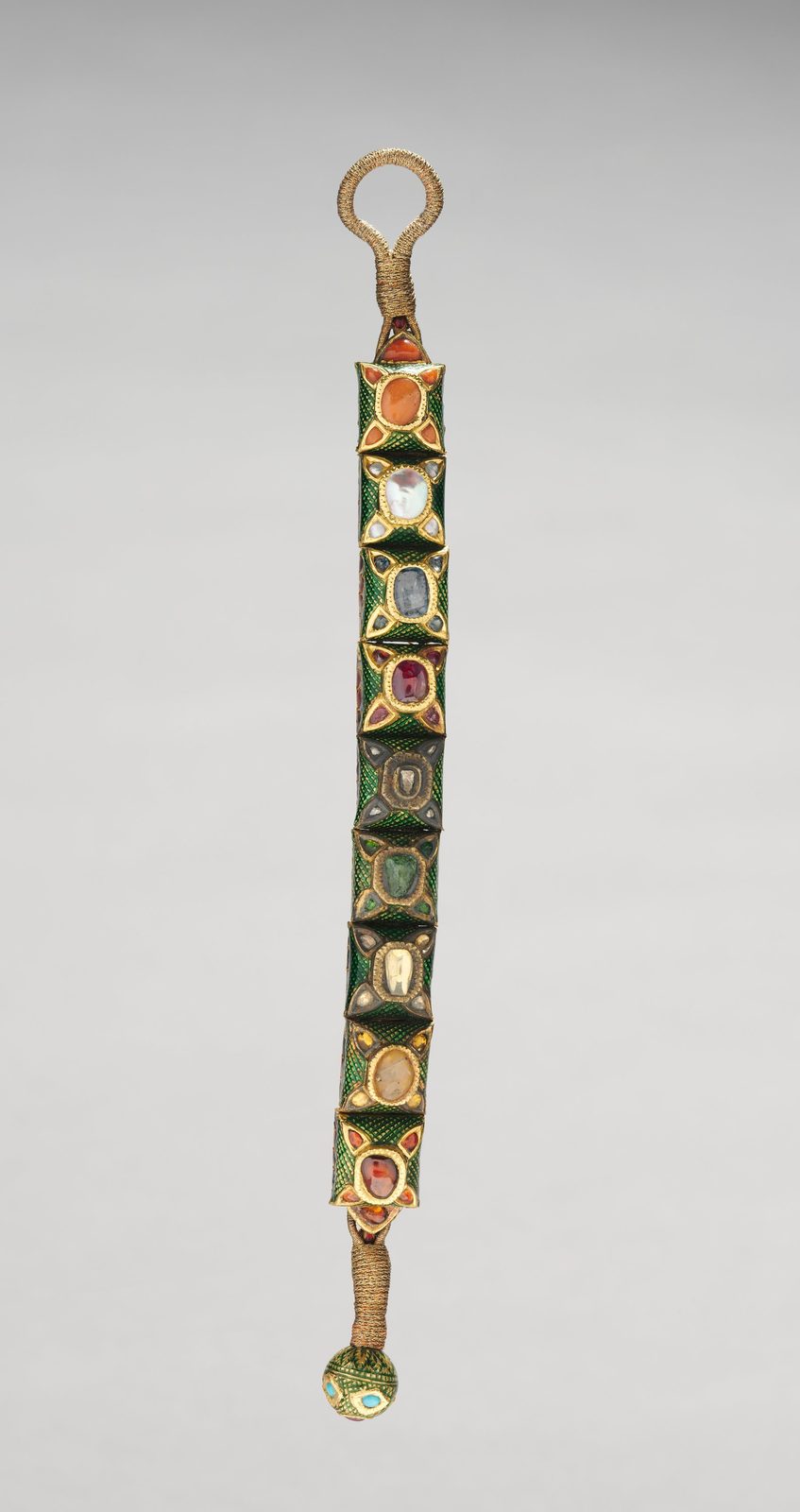 A narrow Indian armband photographed against a plain grey ground. Nine hinged plaques of deep green enamel run down its length, each set in gold with a different stone, orange carnelian, a pearl, a pale blue sapphire, a red ruby, a diamond, a green emerald and others, with a braided gold cord and loop at the top and a small enamelled bead at the bottom.