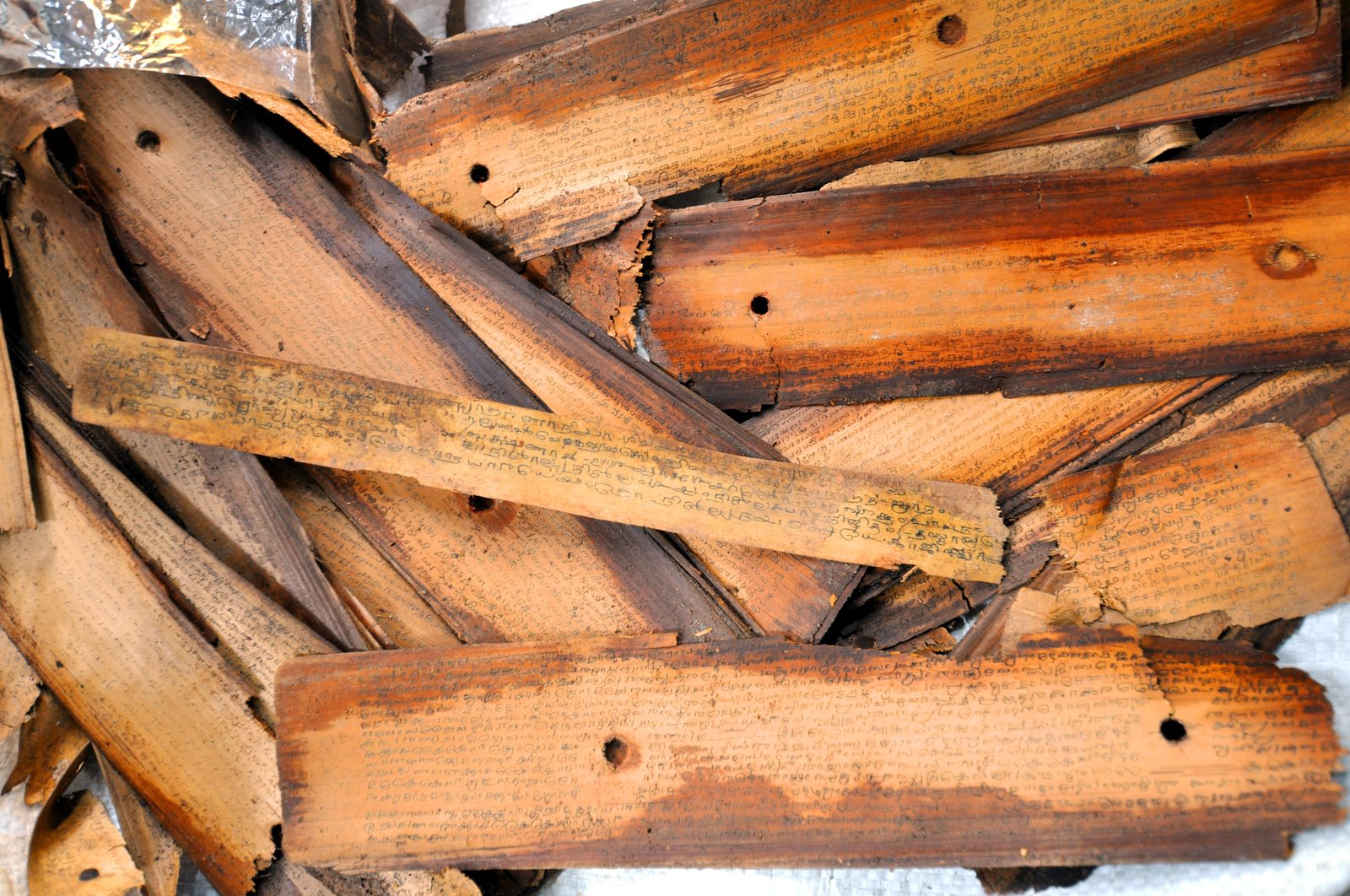 A close pile of loose palm-leaf manuscript strips lying across one another on a white cloth. Each strip is a long narrow blade of dried palm, brown and darkened at the edges, pierced by a round hole for a binding cord, and covered end to end with fine handwritten Tamil letters in dark ink. Several are cracked, chipped or eaten away, and a corner of foil wrapping shows at the top left.