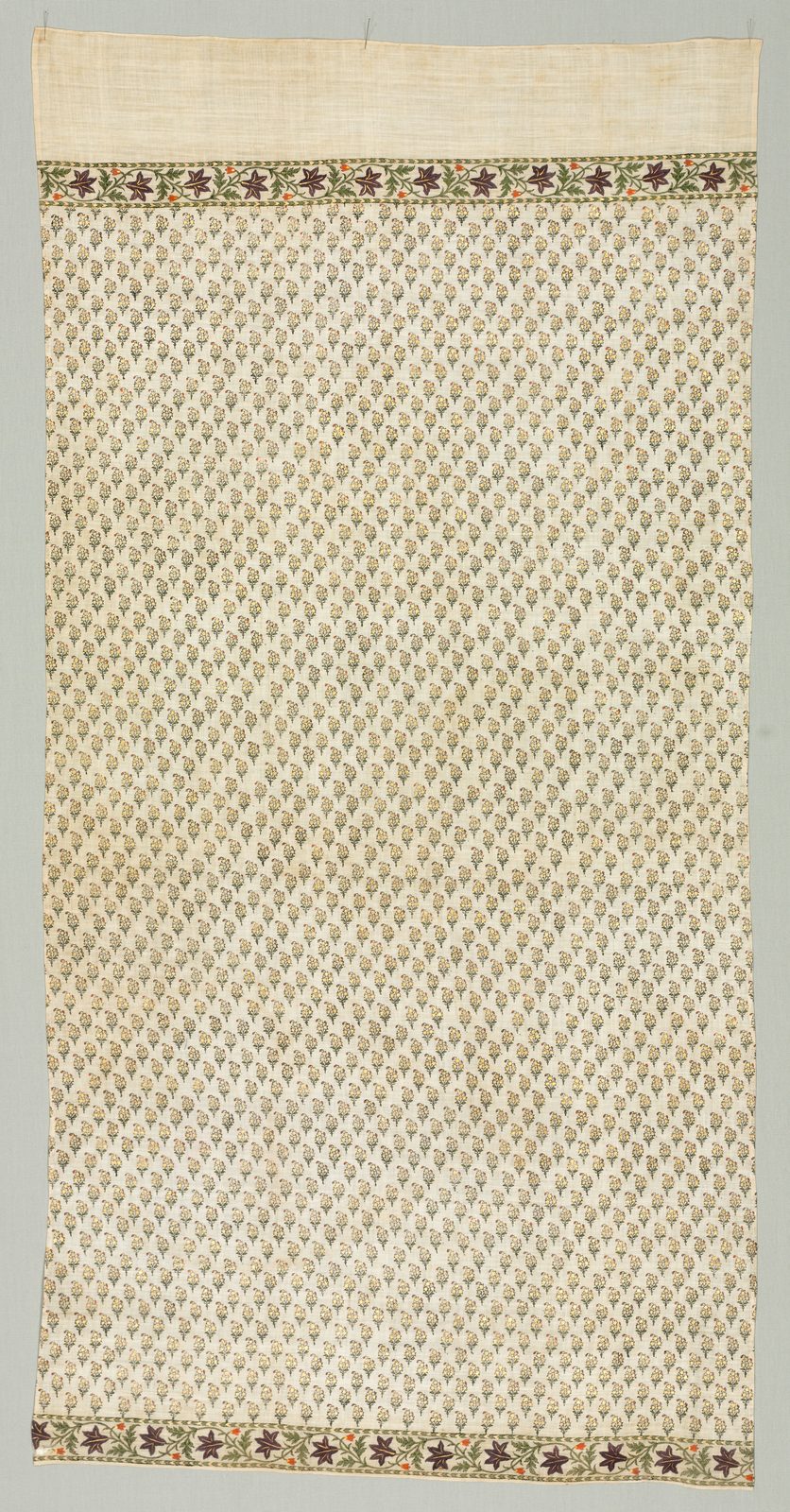A tall cream cotton cloth photographed flat against a pale grey ground. Almost the whole surface is covered with hundreds of small identical printed sprigs, each a tiny leafy spray in faded gold and dark ink, set in even diagonal rows. A narrow band of larger flowers in dark red, green and orange runs across the top and again across the bottom, and a plain undecorated strip of muslin is left along the top edge.
