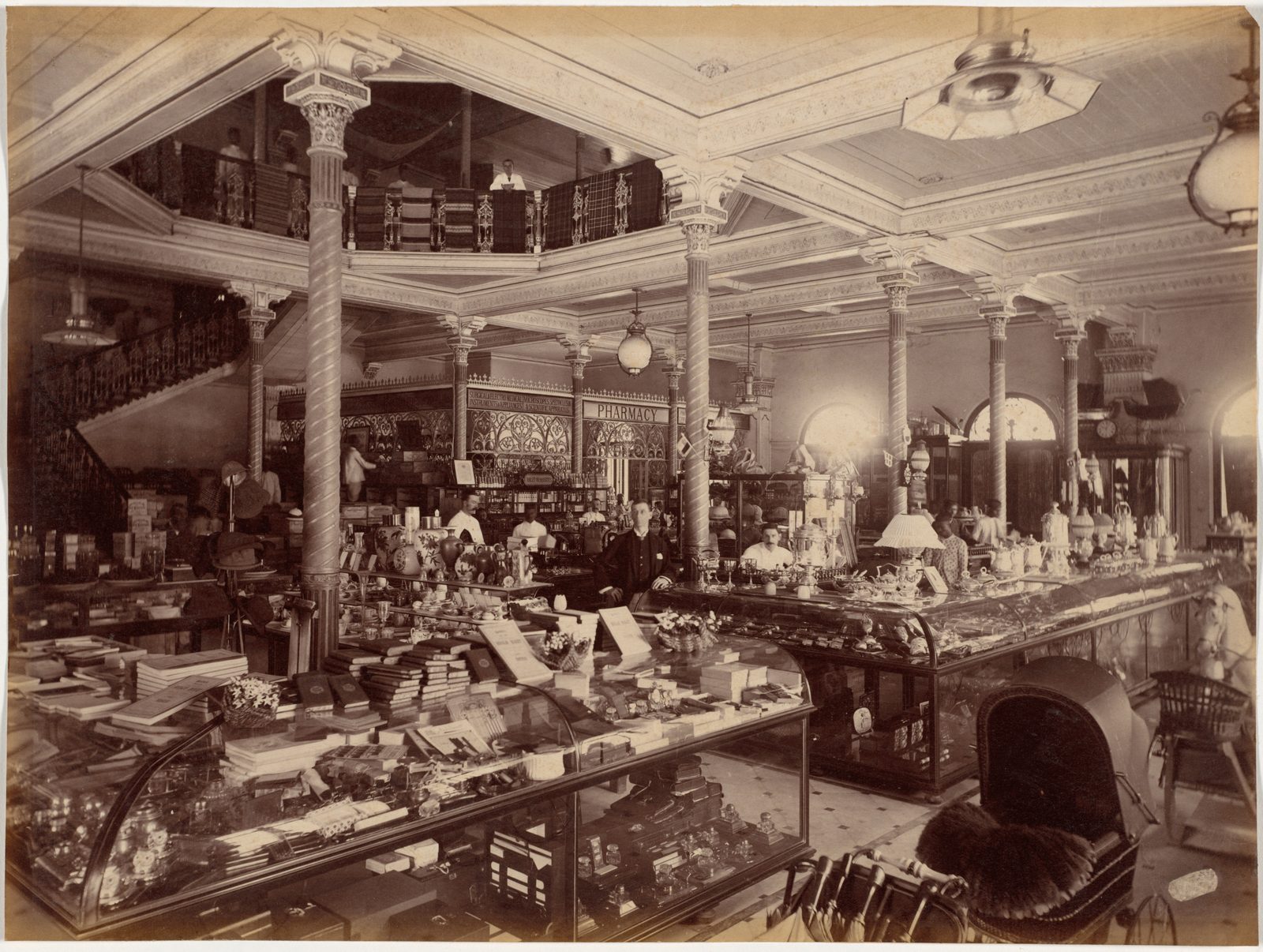 The interior of a large nineteenth century shop in Bombay, photographed from the doorway. Fluted columns run down the room and rise to an upper gallery where figures stand at the rail looking down. Glass-topped counters stacked with books fill the foreground, a baby carriage and a stand of umbrellas stand to one side, and behind further cases of glassware and china the staff wait under a sign for the pharmacy. A patterned ceiling covers the whole room.