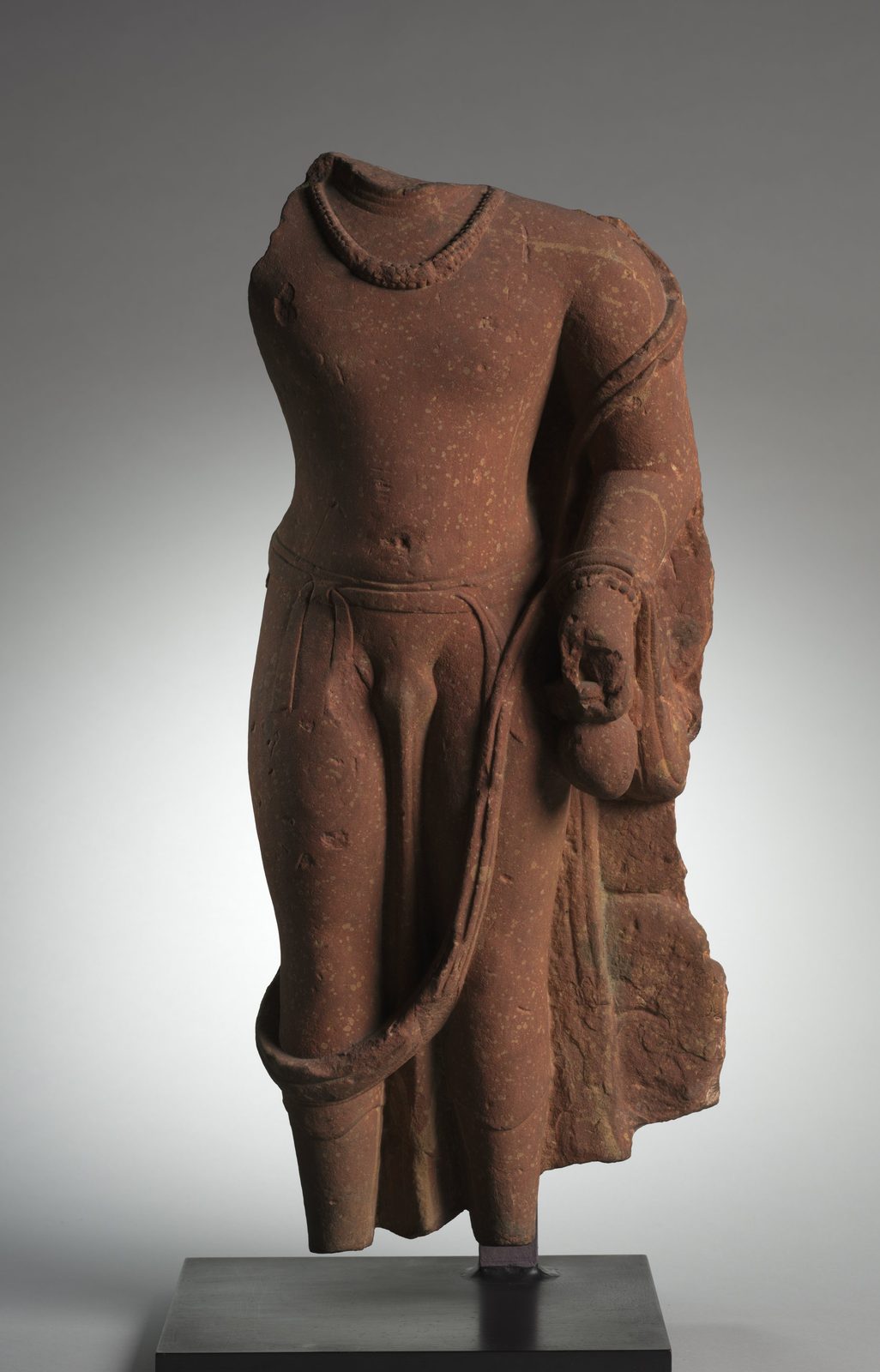 A fragmentary sandstone sculpture of a standing male figure, broken away above the shoulders, below the knees and at the right arm. A beaded necklace lies on the bare chest, a thin garment is gathered at the waist, and the bent left hand holds a small round water pot. The body of a serpent is draped over the left shoulder and loops across the front of the thighs.