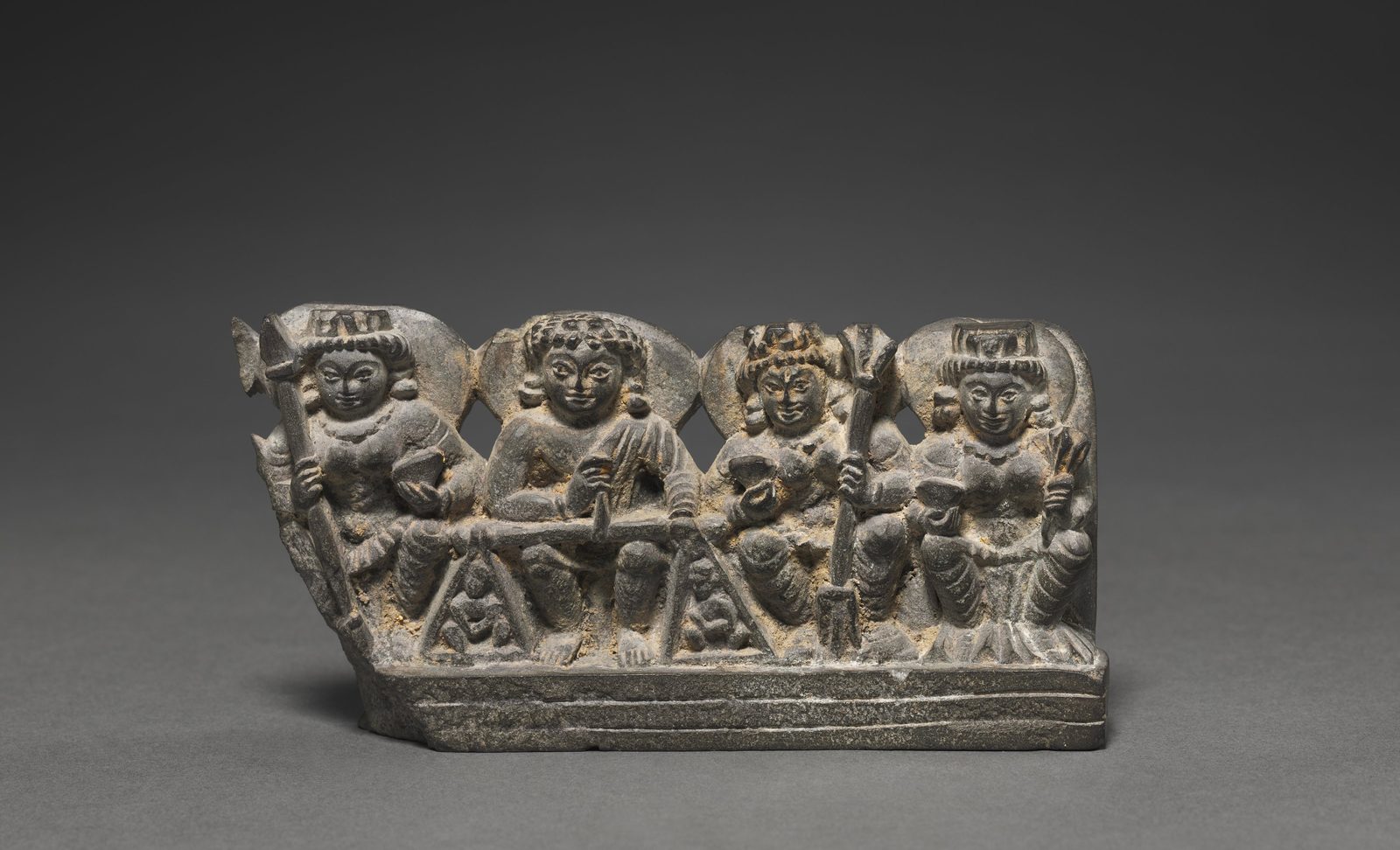 A small dark schist relief carving of four seated figures in a row, each haloed and crowned. The central figure, Yama, h