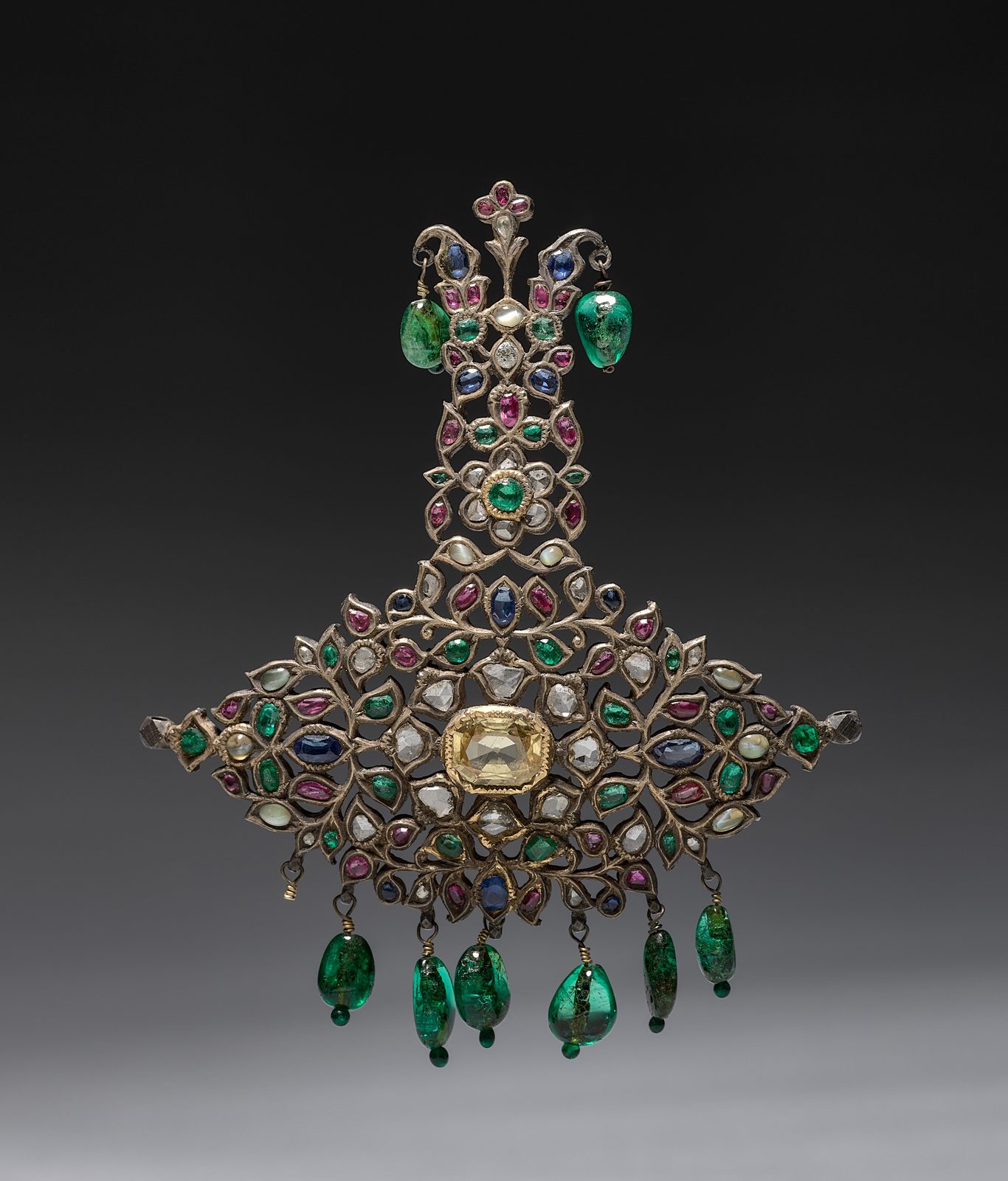 A small Indian turban ornament photographed against a graded grey ground. A wide fan-shaped base of openwork silver is set all over with flat-cut green, red, blue and colourless stones in leaf-shaped mounts, with a large pale yellow rectangular stone at its centre. An upright plume rises from the fan, narrow at the foot and scrolling open at the top, with a green teardrop bead hanging on either side of it. A row of green teardrop beads hangs along the lower edge of the fan.