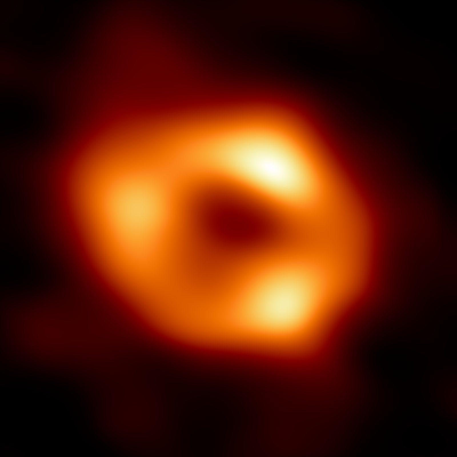 A soft, blurred ring of orange and yellow light on a black field, brightest at three uneven patches spaced around its rim, with a dark rounded hollow at the centre and a faint red halo outside it. There is no other detail in the frame.