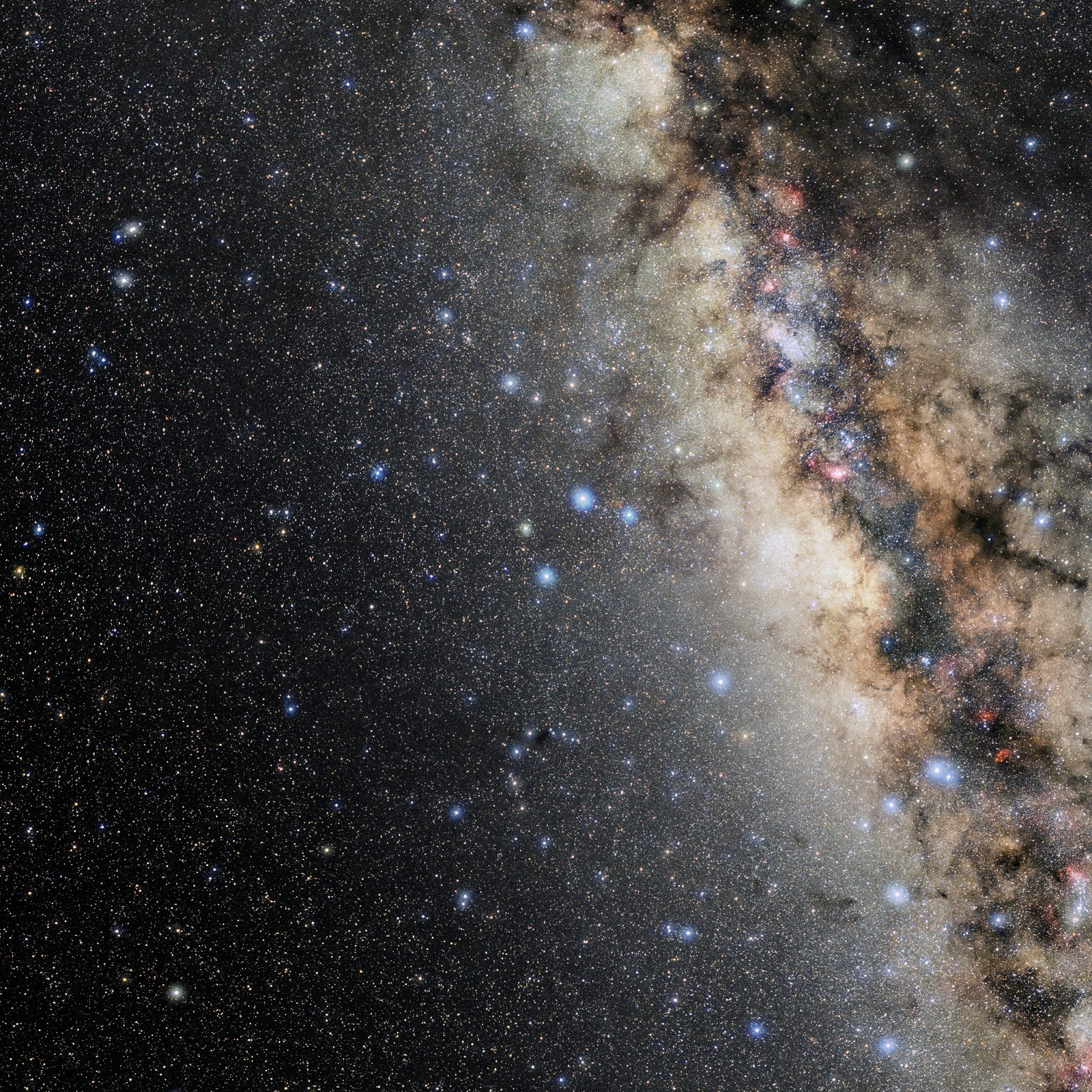 A wide colour photograph of a star field. The left half is nearly black and scattered with faint pinpoints; the right half is filled by a dense pale band of unresolved stars, warm cream and tan in tone, crossed by ragged dark lanes of dust and dotted with small pink patches. A scattering of bright blue-white stars stands in front of the band.