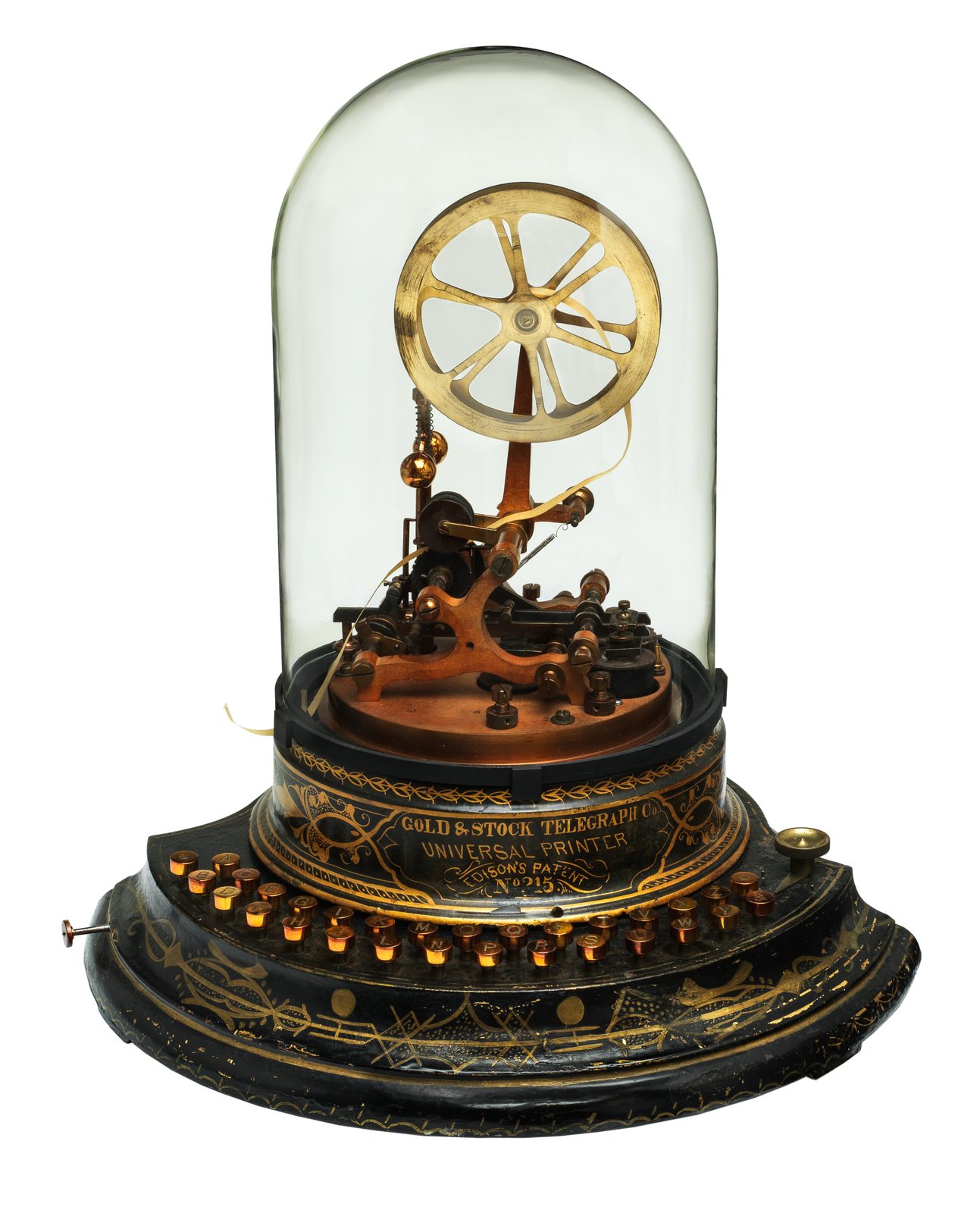 A museum photograph of a nineteenth-century stock ticker standing alone on a plain white ground, taller than it is wide. The machine sits on a wide black wooden base with two stepped tiers, painted with faded gold scrollwork. Around the upper tier runs a curved row of about thirty small round brass keys, each with a letter of the alphabet on it. Above the keys a black drum carries gold lettering in three lines: GOLD & STOCK TELEGRAPH Co., then UNIVERSAL PRINTER, then EDISON'S PATENT, with a number beneath. A tall clear glass dome covers the working parts: a large spoked brass flywheel standing upright at the top, a brass and copper mechanism of levers, screws and coils below it, and a narrow paper tape that feeds through the printing head and curls out from under the rim of the dome onto the base. No person is in the picture and nothing is printed on the tape.