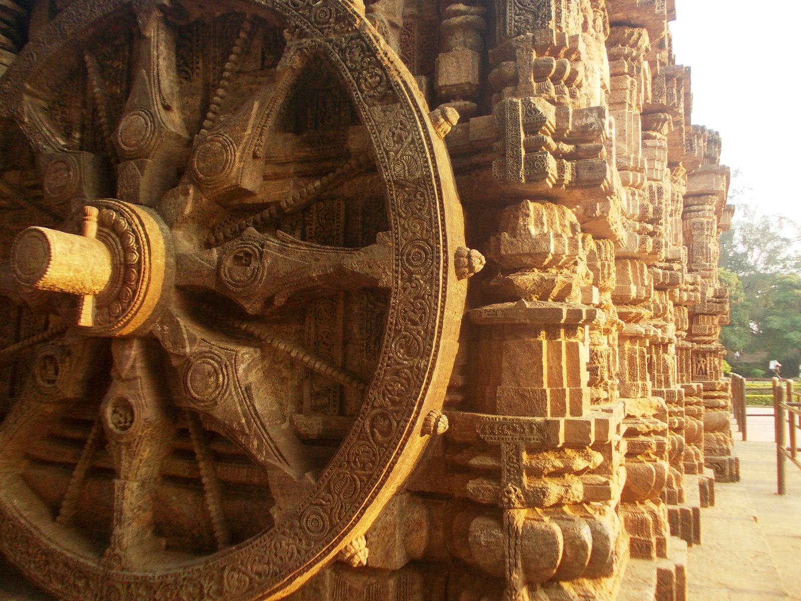 A huge carved stone chariot wheel fills the left of the frame, seen at a sharp angle in warm late light. Its thick spokes carry round carved medallions, a decorated axle projects from the hub, and the broad rim is cut with foliate ornament and a beaded band. To the right the temple's carved sandstone mouldings step away into the distance, with a low railing, trees and pale sky beyond.