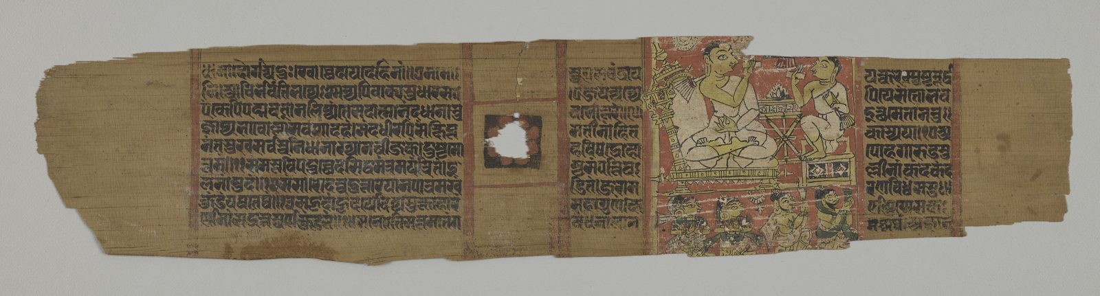 A long, narrow palm-leaf manuscript folio photographed flat against a pale grey ground. Most of the leaf carries close-set lines of Sanskrit in an early Devanagari hand, divided into blocks by red rules, with a small red-framed blank square set into the text left of centre where the string hole was reserved. At the right end a painted panel on a red ground shows a seated teacher in white raising one hand towards a smaller seated pupil, a manuscript resting on a low stand between them, and a row of lay listeners along the bottom. Both ends of the leaf are chipped.