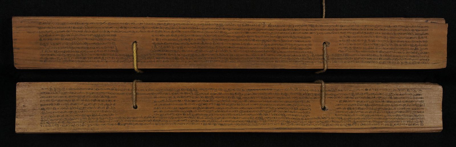 Two long, narrow palm-leaf manuscript folios laid one above the other and photographed against a black ground. Each pale brown leaf is closely written in five or six lines of small Malayalam script in dark ink, with two round string holes punched through it and a loop of pale cord still threaded through the upper leaf. The edges are worn and chipped.